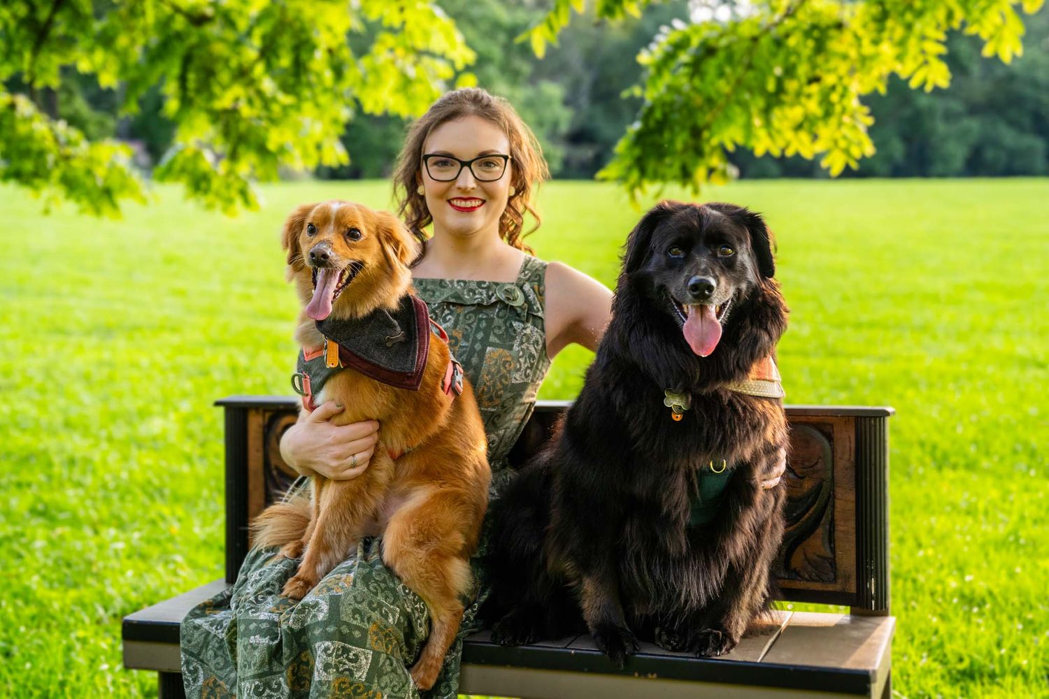 Smiling woman sitting on bench with golden and black fluffy dogs in a green park.