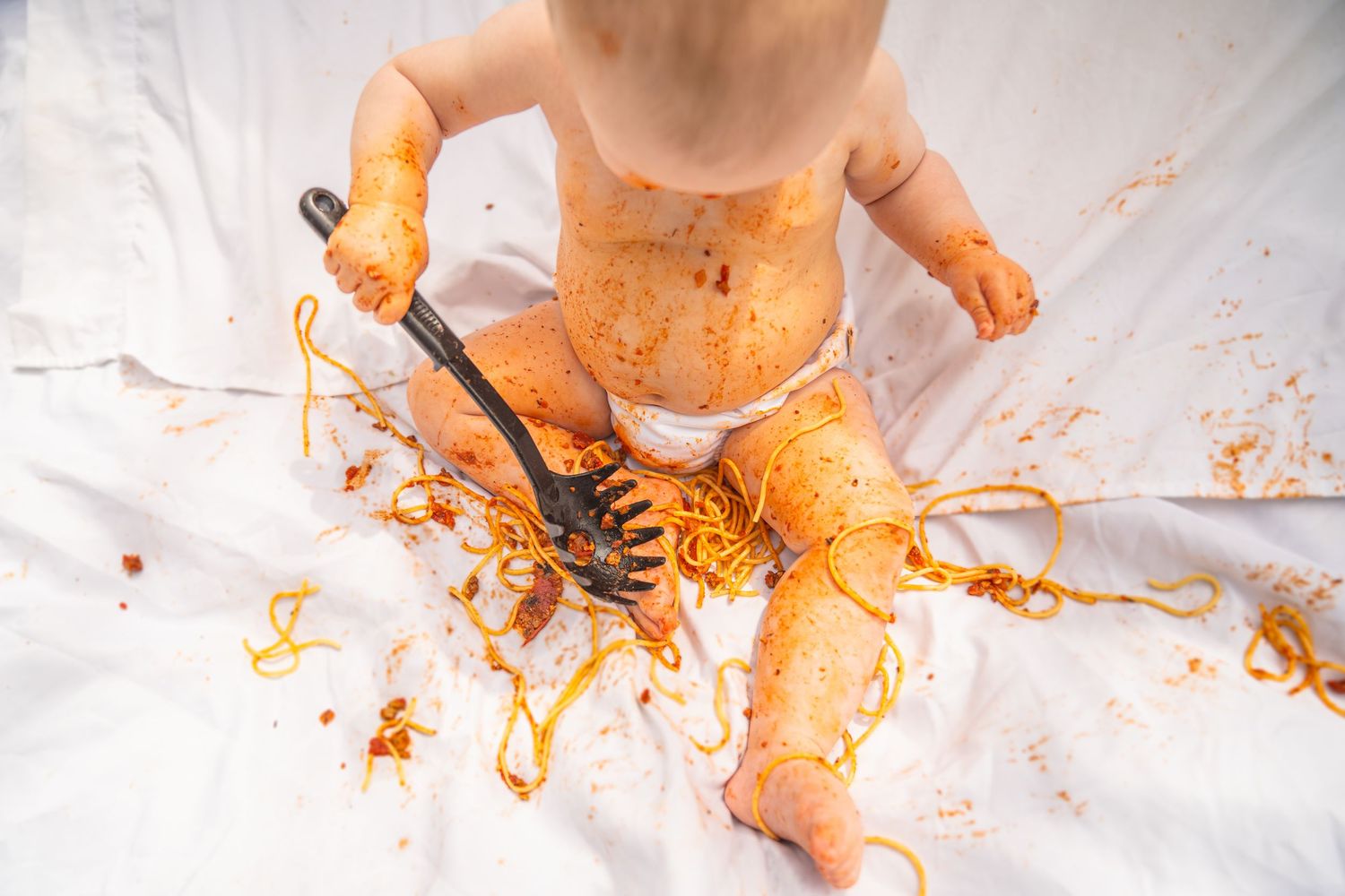 Baby in diaper covered in spaghetti sauce, holding a fork with pasta, sitting on a white sheet.