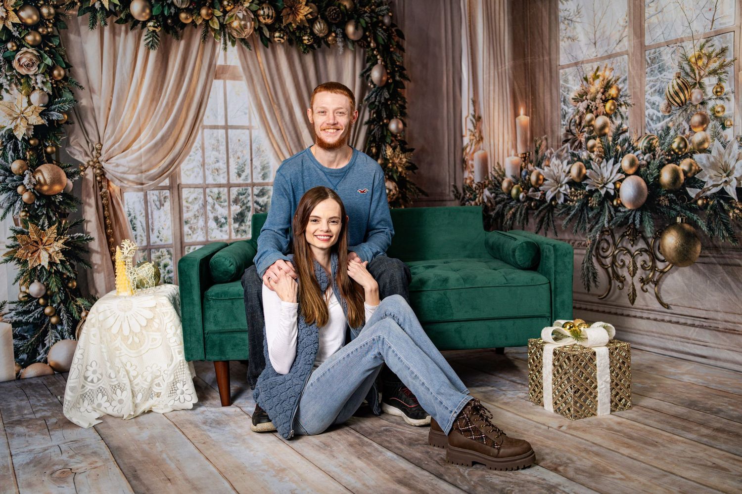 Couple posing in festive Christmas studio with green velvet sofa, decorated tree, and gifts on wooden floor.