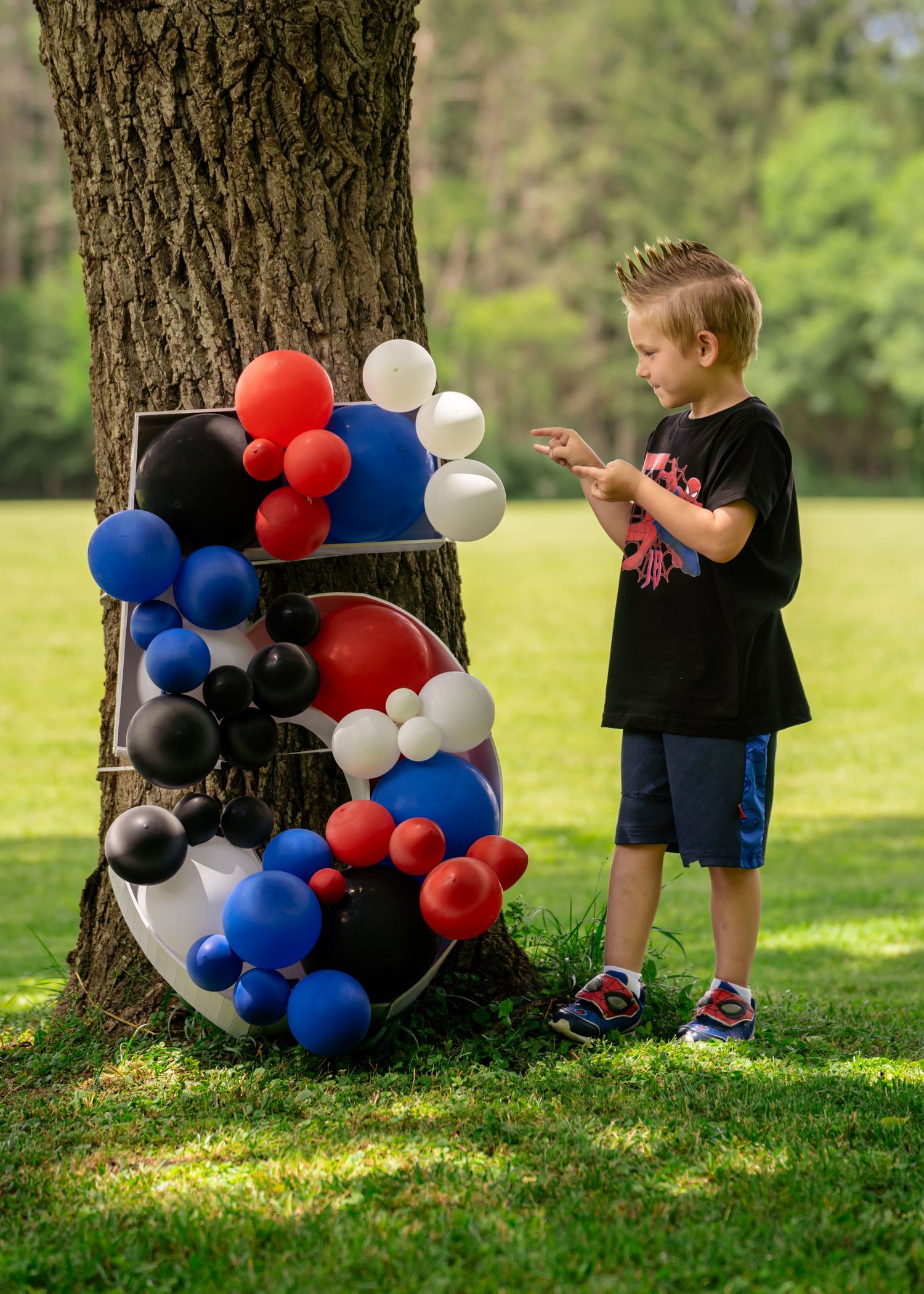 A young boy stands beside a number 5 balloon sculpture with red, blue, black, and white balloons near a tree outdoors.