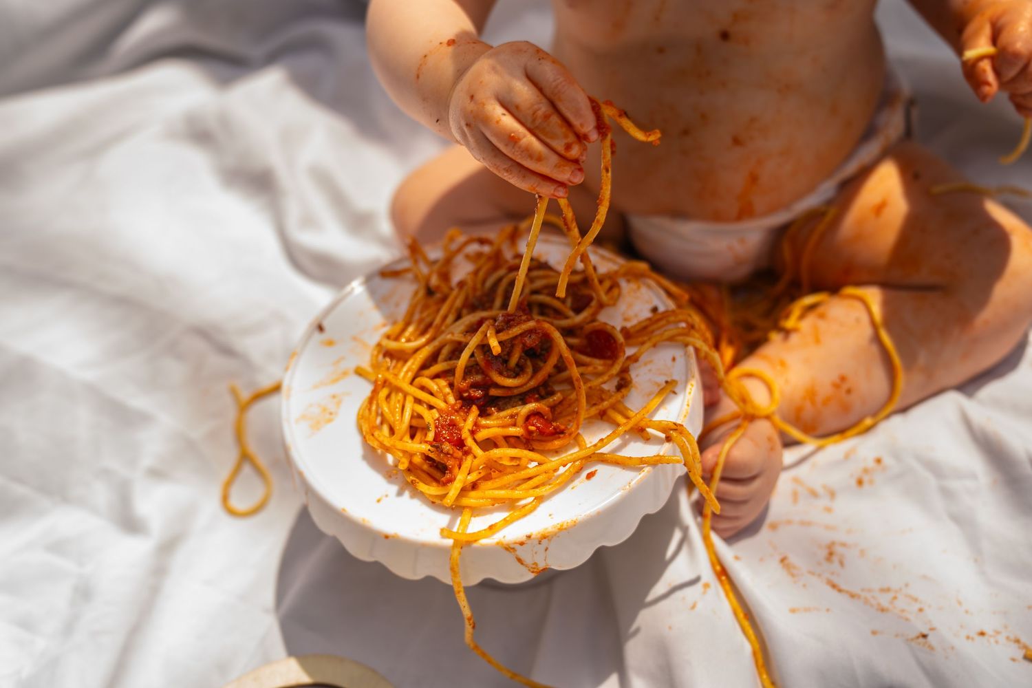 Baby in diaper joyfully playing with spaghetti in tomato sauce on a white plate, hands and body covered in mess.