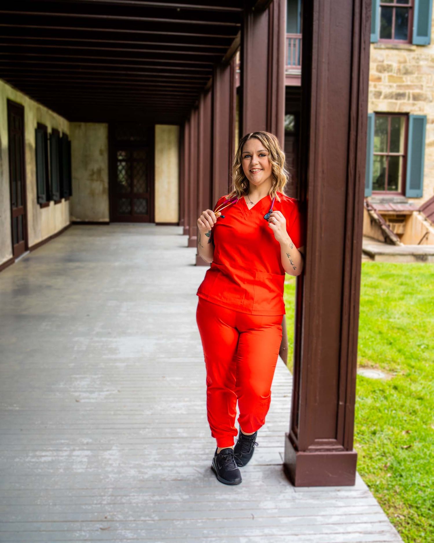 Smiling woman in red scrubs walks along covered outdoor hospital corridor.