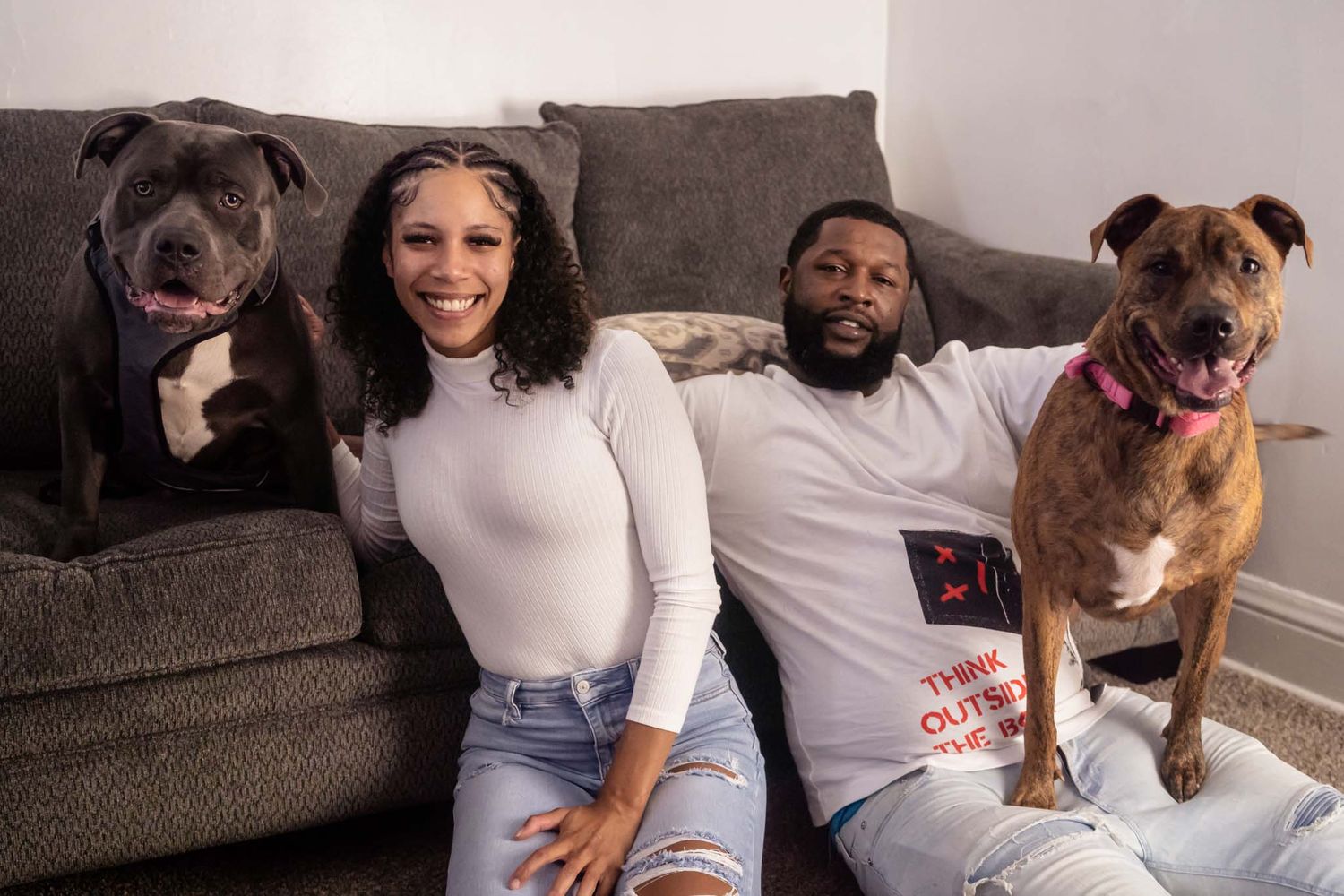 Smiling couple sitting on floor with two pit bull dogs beside a gray couch in a cozy living room.