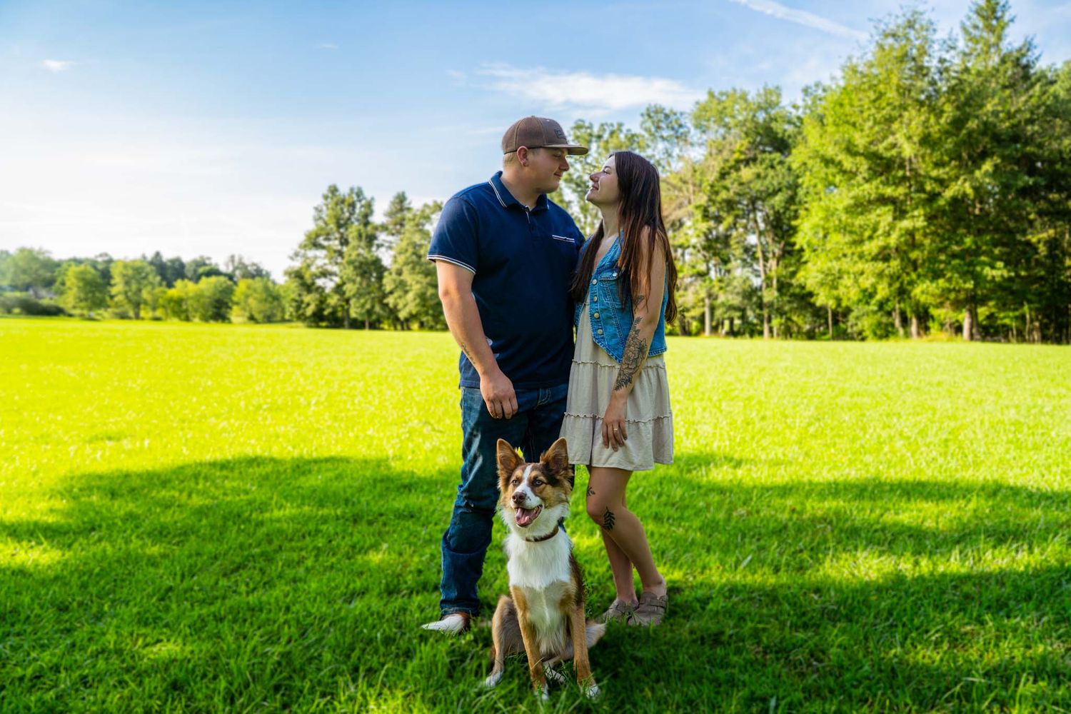Couple stands together smiling in sunny green field while brown and white dog sits at their feet.