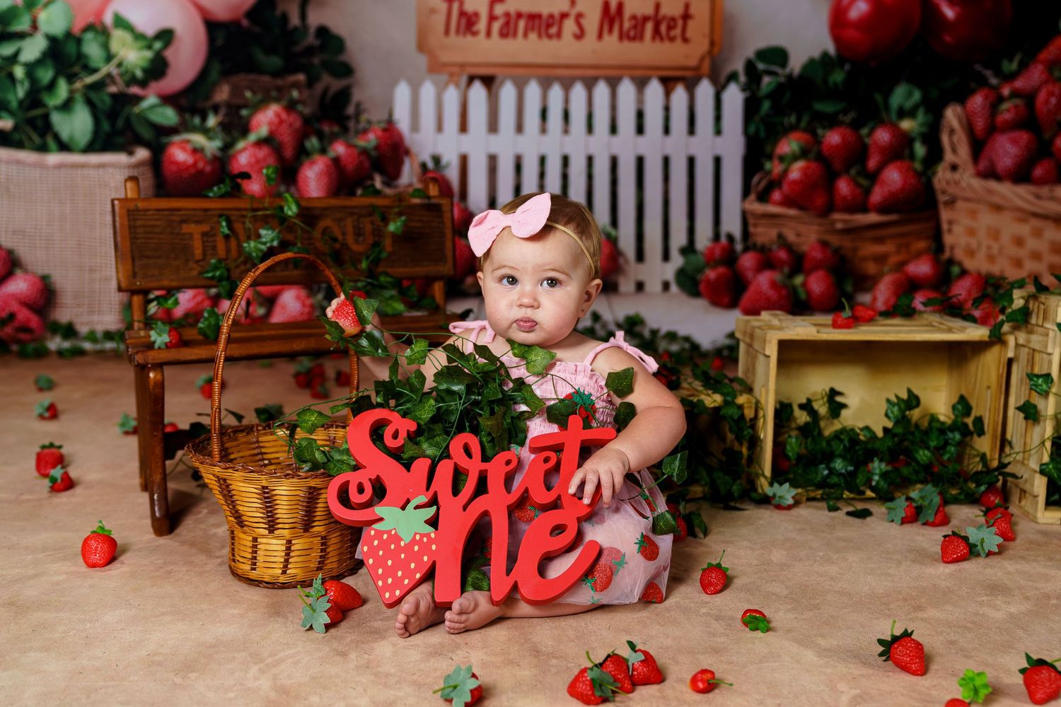 Adorable baby girl in pink dress sitting at a strawberry-themed first birthday cake smash setup.