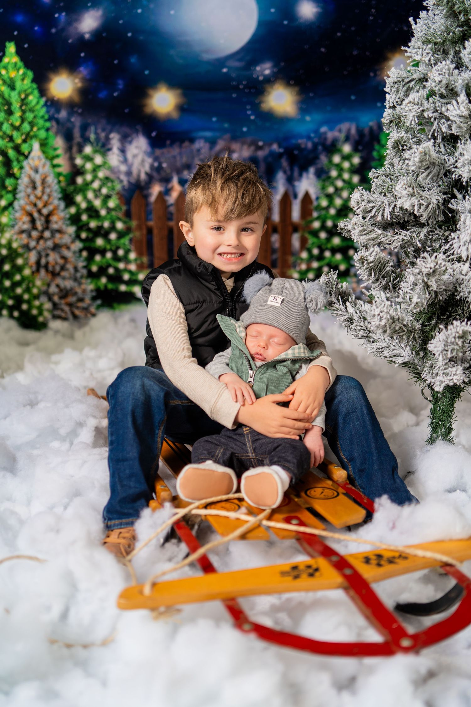 Smiling boy holding a sleeping newborn on a sled in a festive Christmas winter scene backdrop.