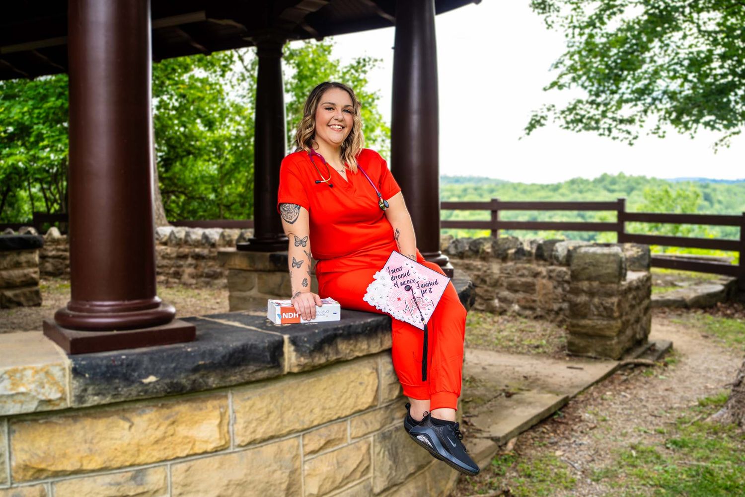 Woman in red scrubs sits at stone pavilion overlooking scenic landscape.