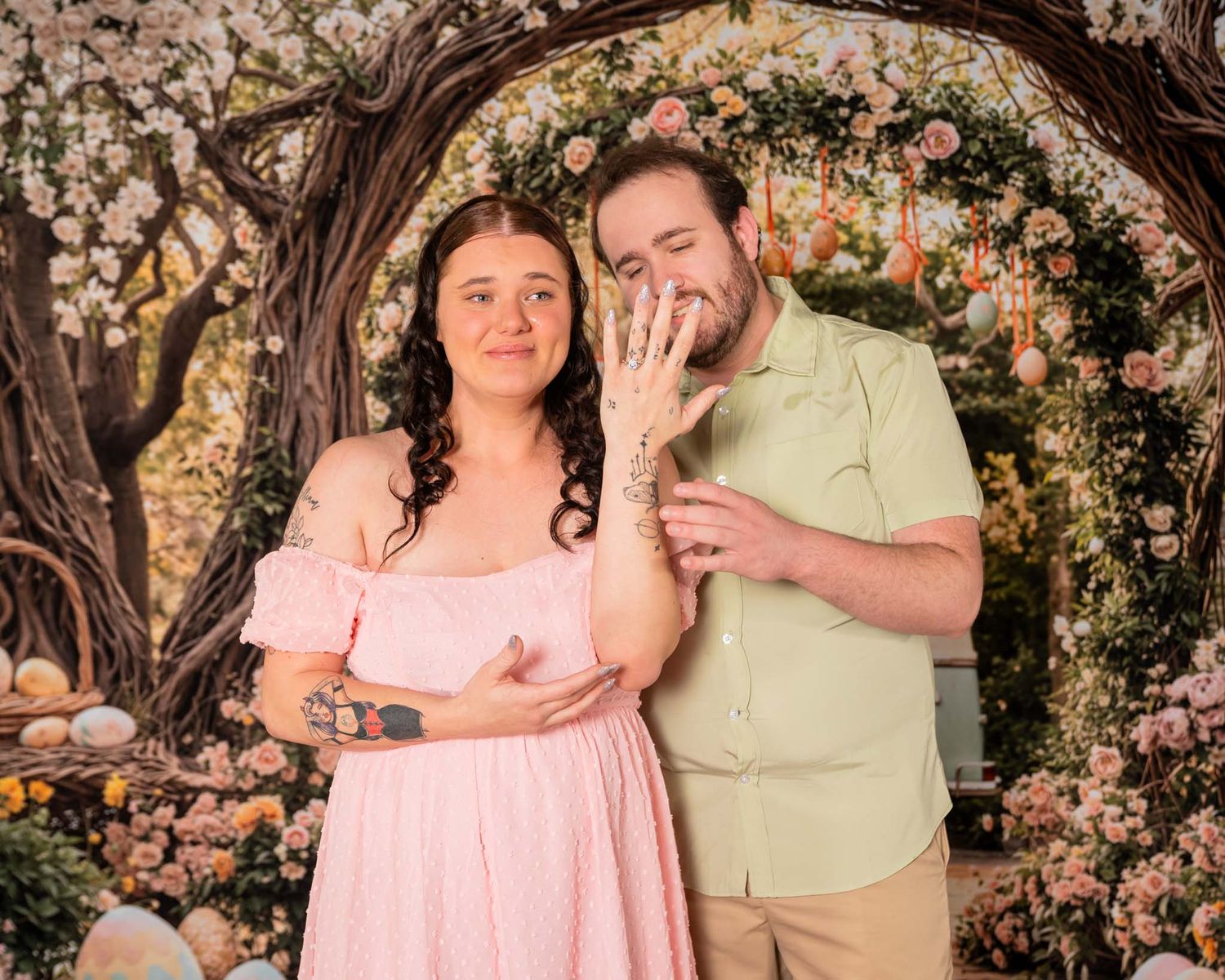 Couple poses under floral arch, woman in pink dress showing engagement ring, man in green shirt, surrounded by flowers.