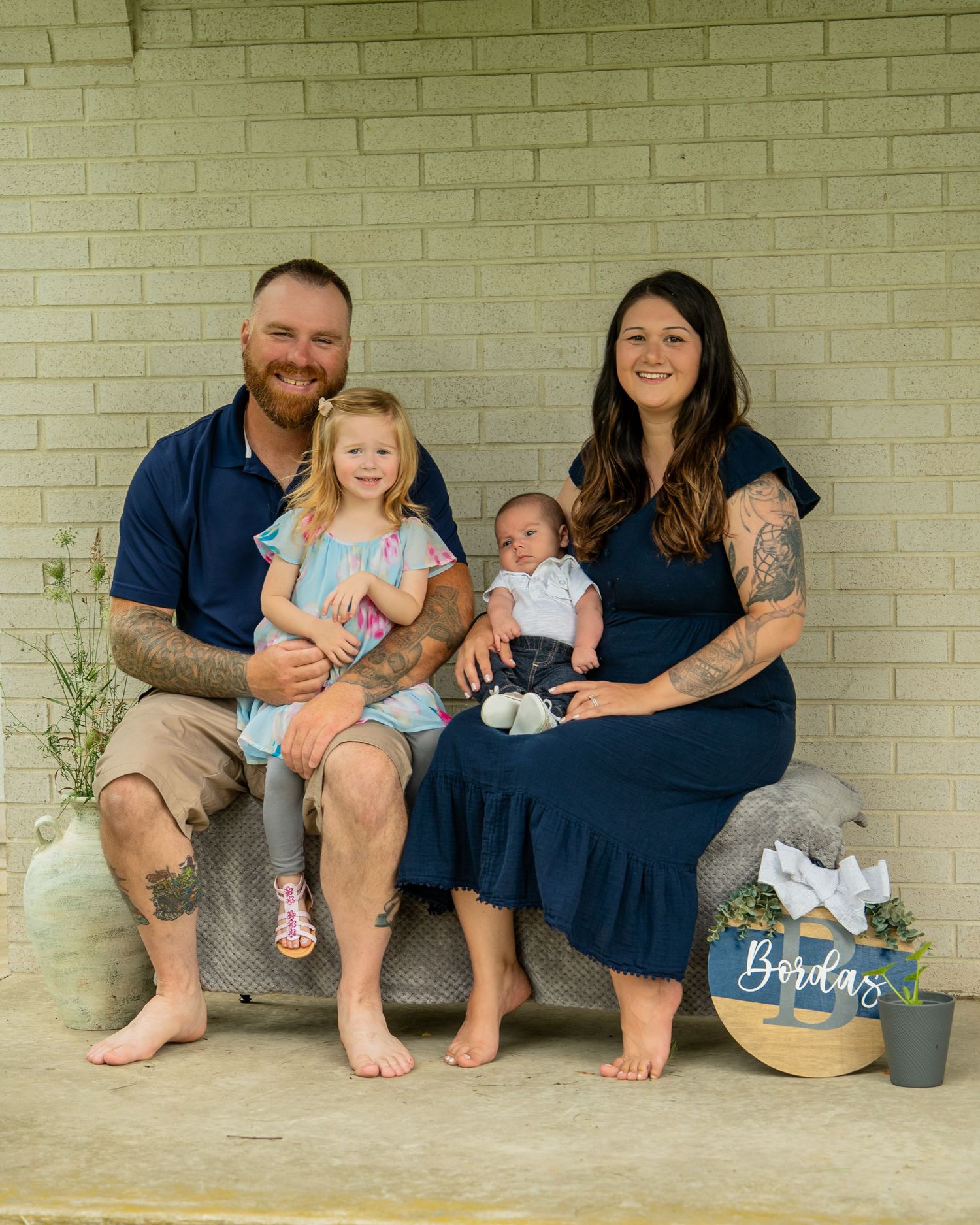 Family of four posing outdoors, parents holding two young children, sitting near a Bordas sign with greenery.