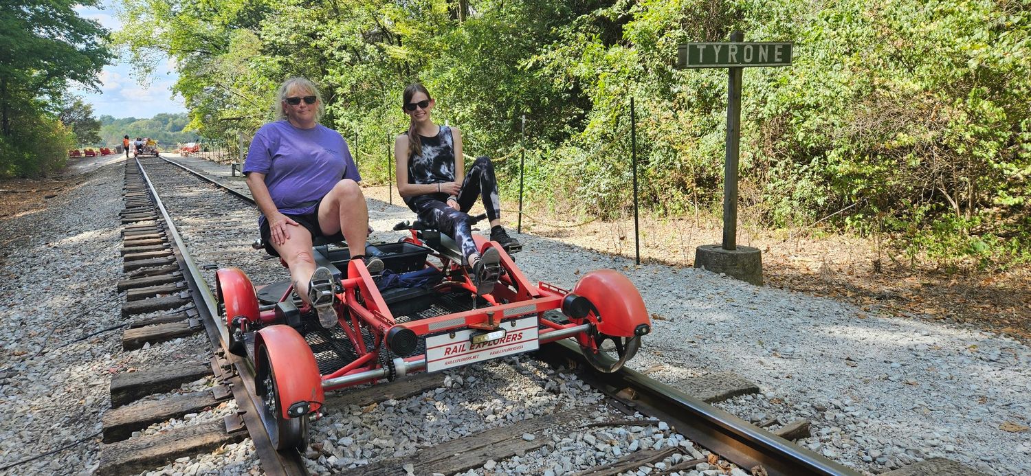 Two people riding a red rail bike on a scenic wooded railway track on a sunny day.