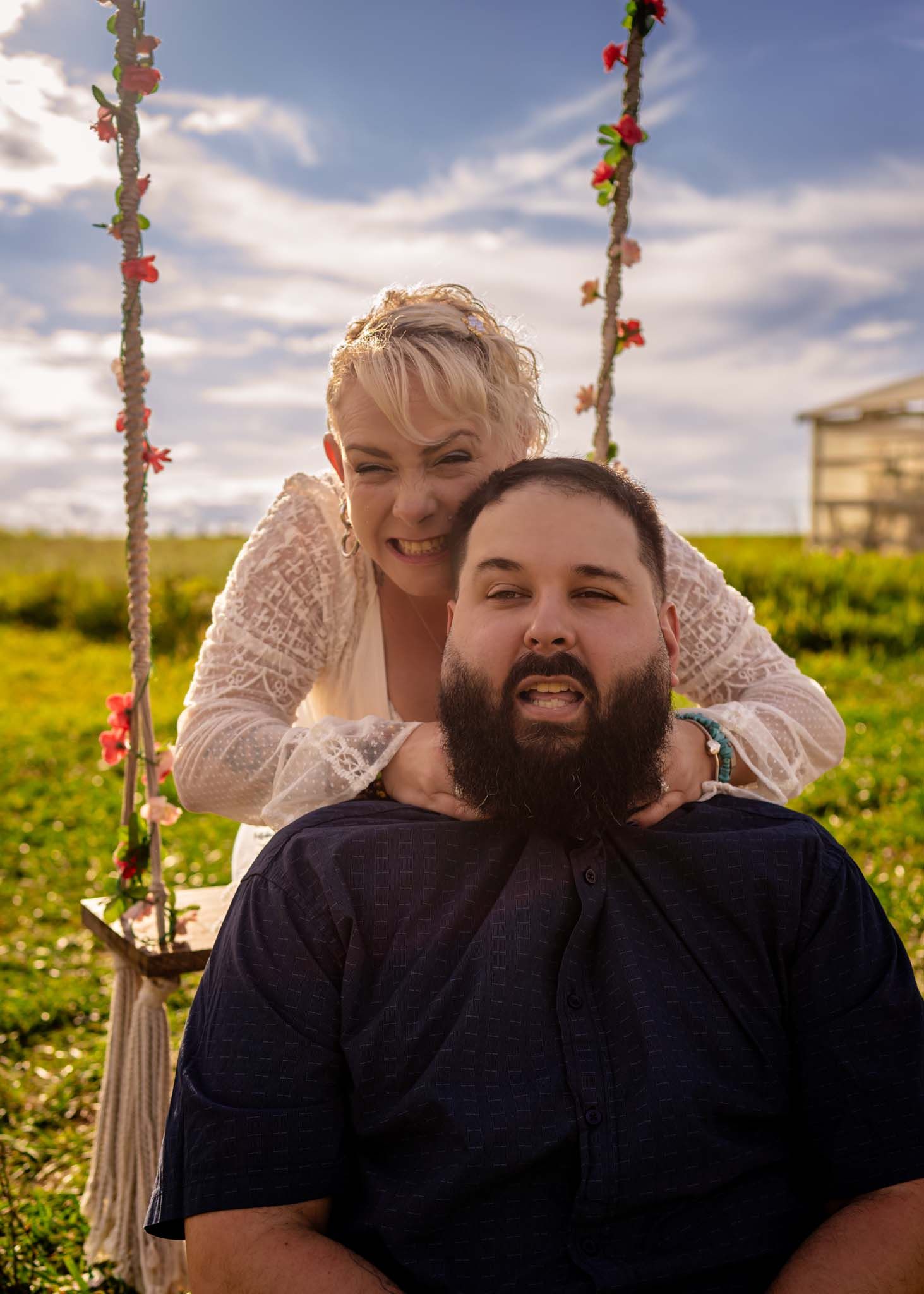 Couple posing outdoors on a floral swing, woman standing behind bearded man, green field and cloudy sky background.