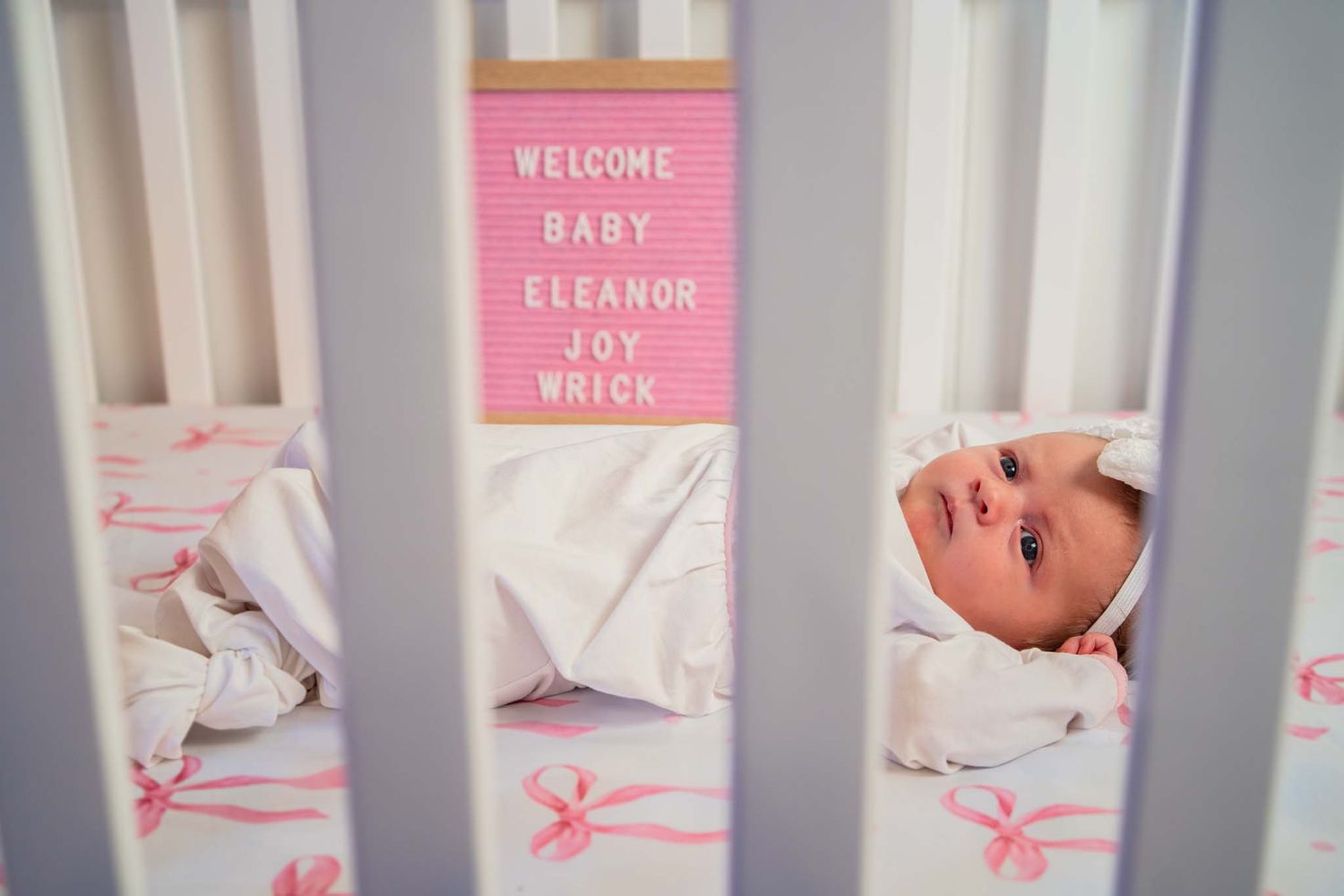 Newborn baby in white crib with welcome sign reading Baby Eleanor Joy Wnick.