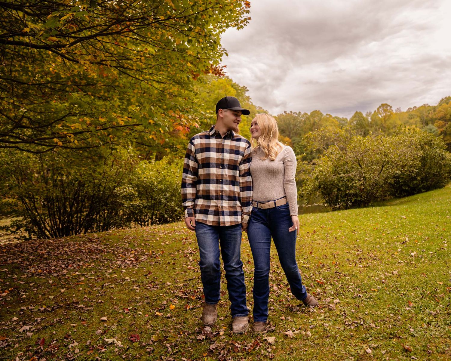 A couple walks together through a lush green park during autumn, surrounded by colorful fall foliage.