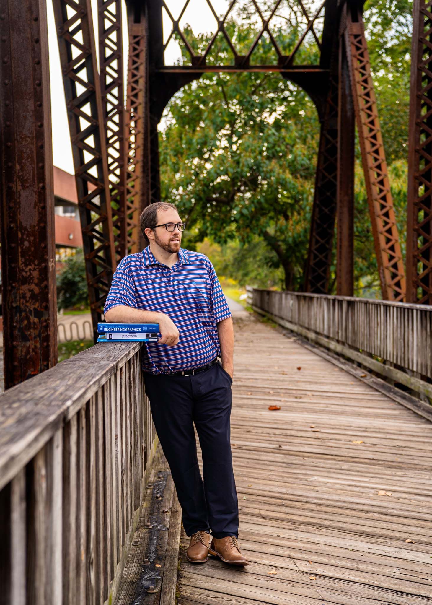 Man in striped polo shirt leaning on railing of rustic iron bridge with books.