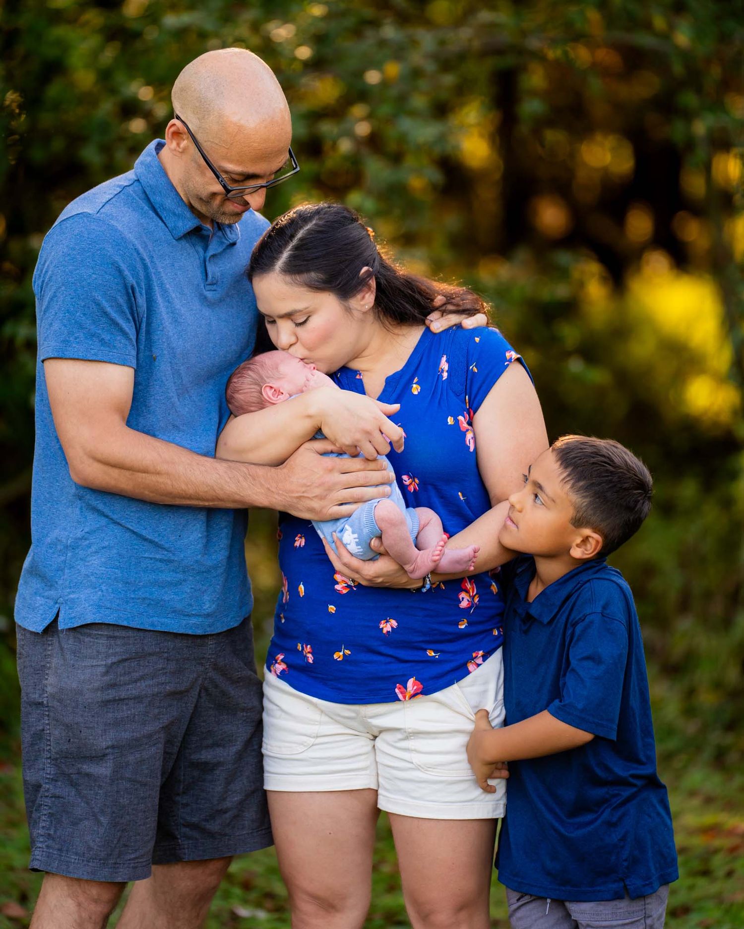 Family of four with parents in blue shirts kissing newborn baby while young son looks on.