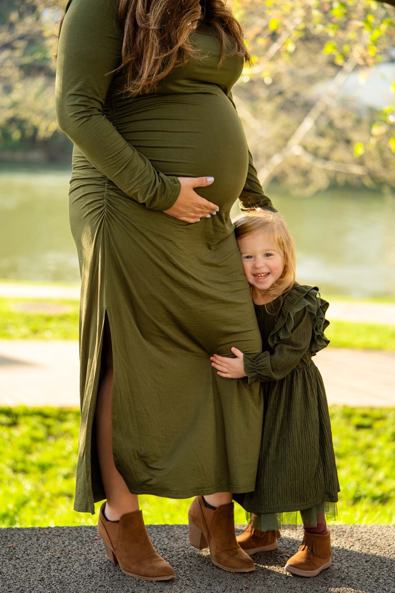 A pregnant woman in an olive green dress stands outdoors as a toddler hugs her belly.