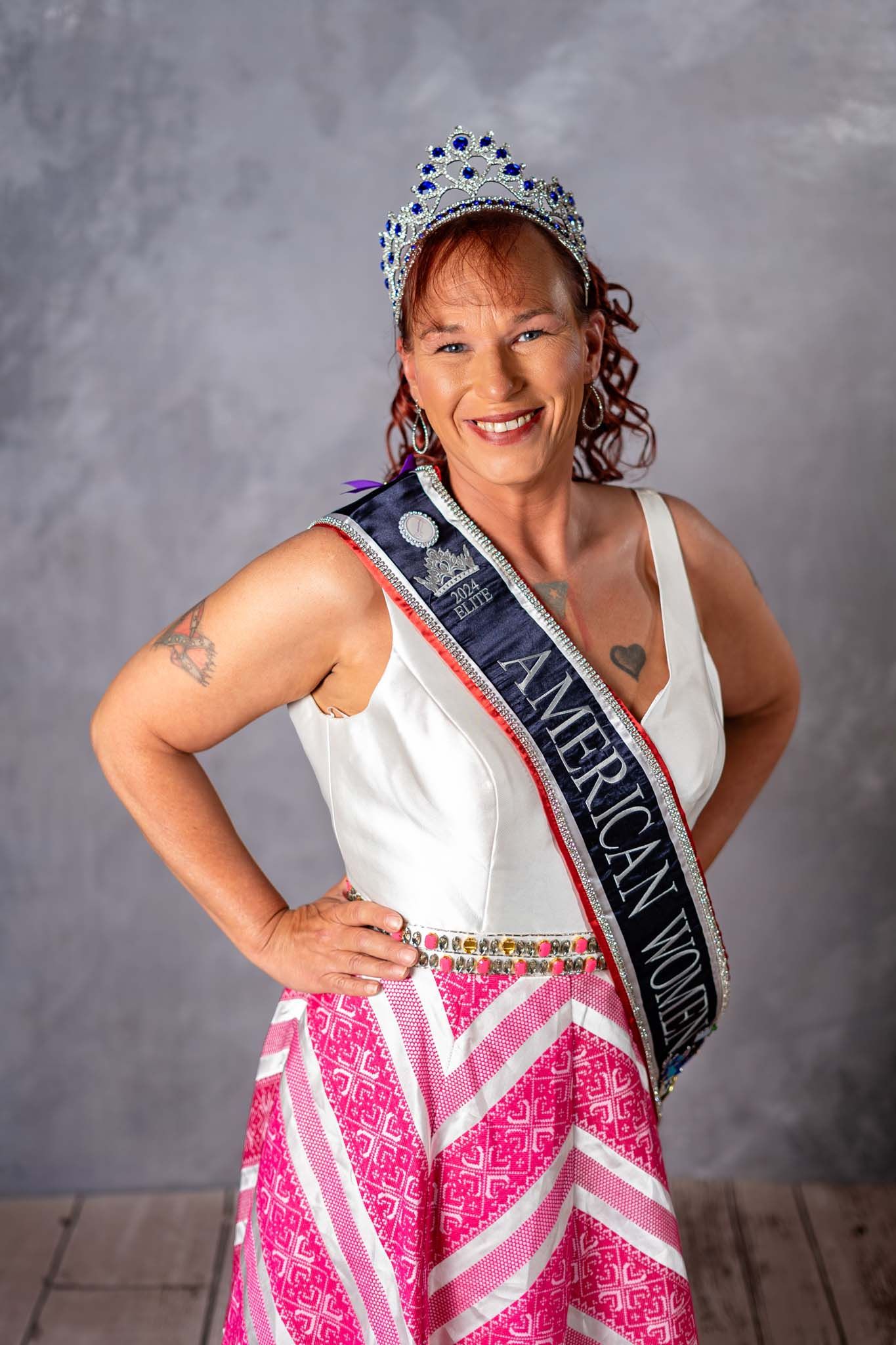 Woman wearing American Women pageant sash and jeweled crown in white top and pink skirt posing confidently.