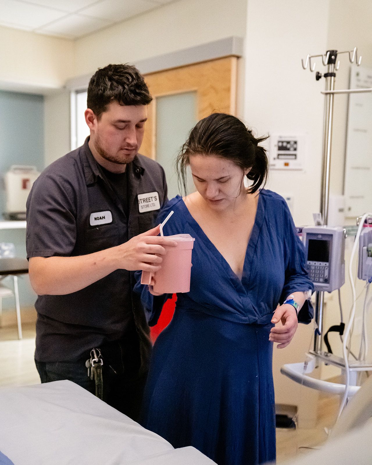 Supportive partner helping laboring woman walk in hospital room while she drinks from a cup.