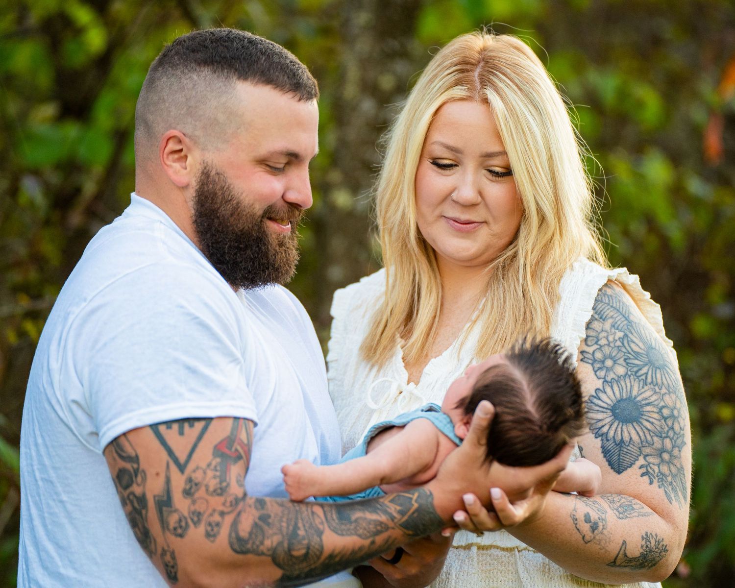 Tattooed couple tenderly holding their newborn baby outdoors with green foliage in the background.