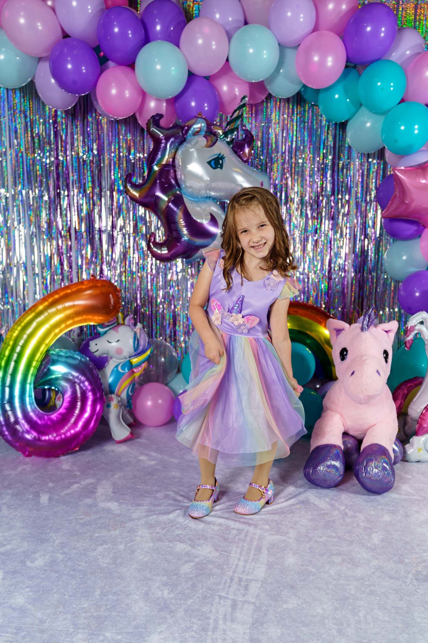 A girl in a purple unicorn dress poses at a colorful 6th birthday party with balloons and stuffed animals.
