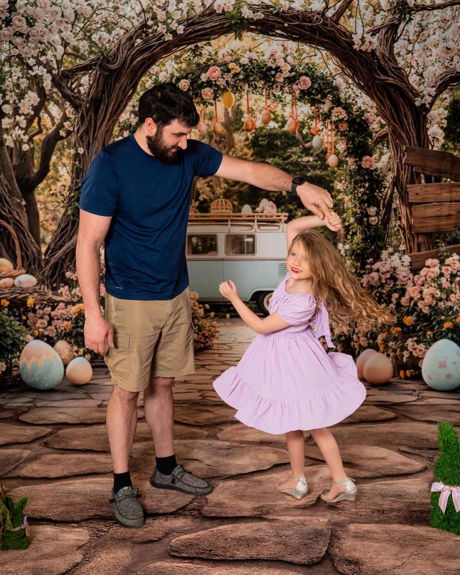 A father twirls his young daughter in a lavender dress under a floral arch with Easter decorations outdoors.