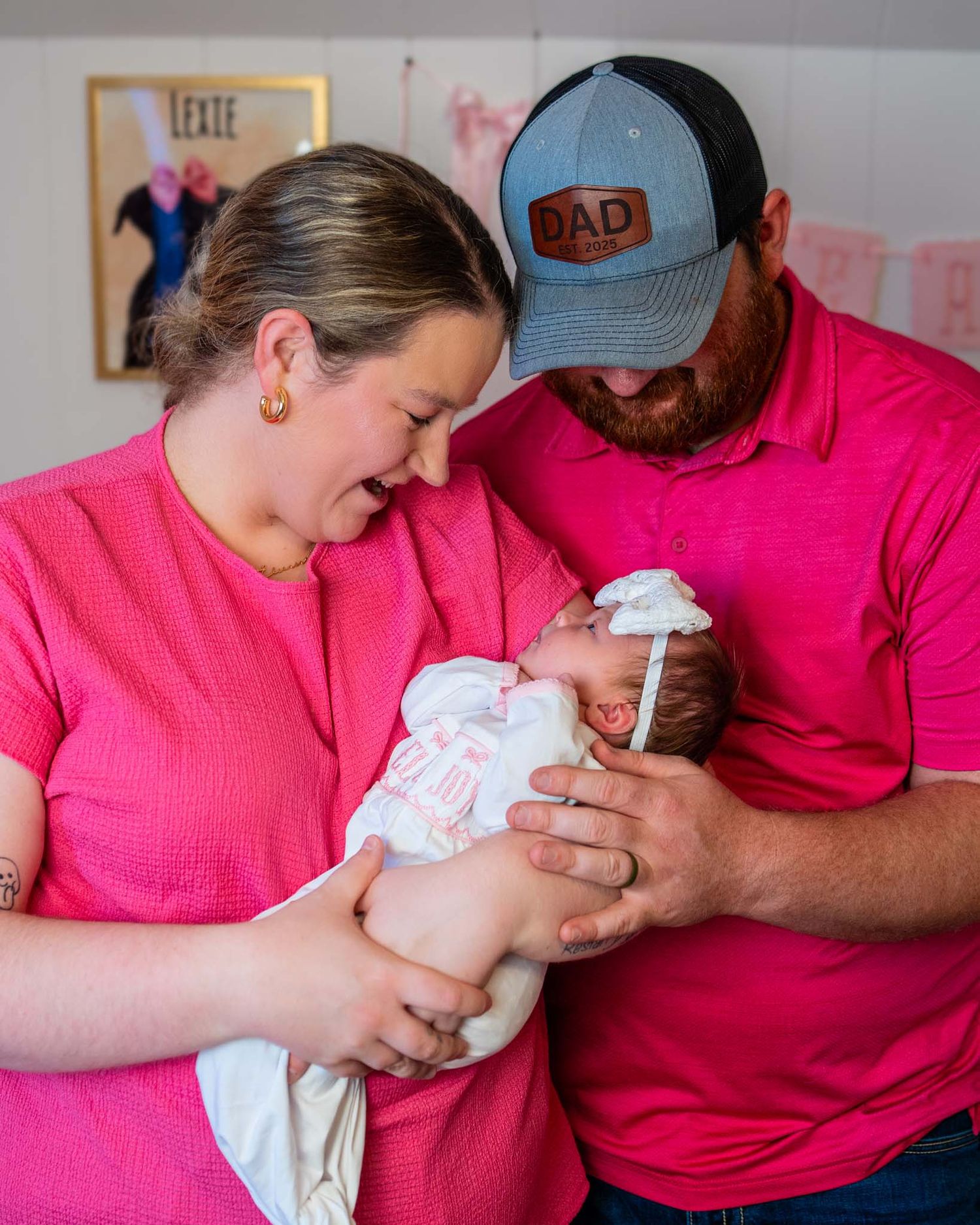 New parents in matching pink shirts lovingly hold their newborn baby in a decorated nursery room.