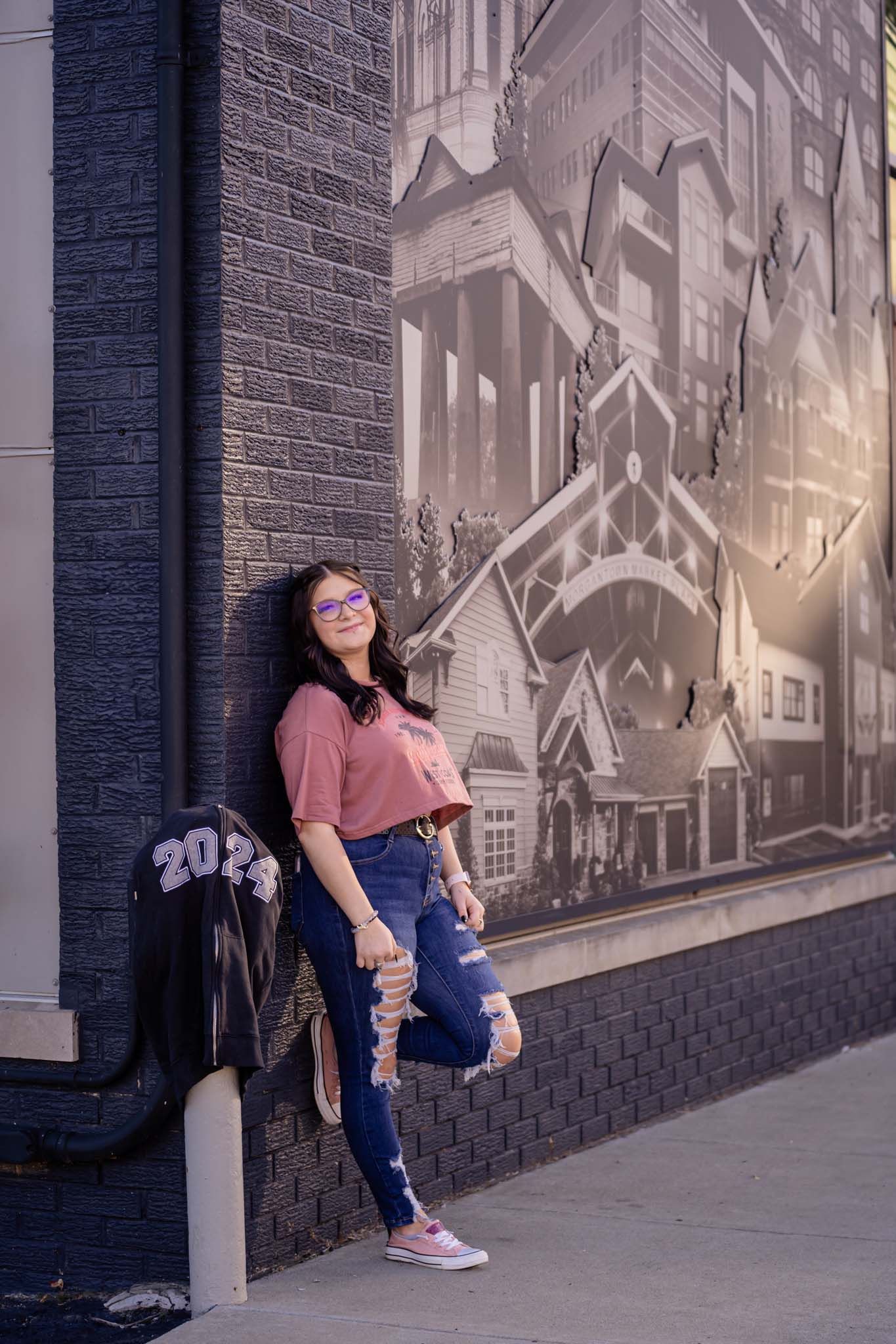 Young woman in ripped jeans and pink top leaning against a brick wall mural for 2023 graduation photos.