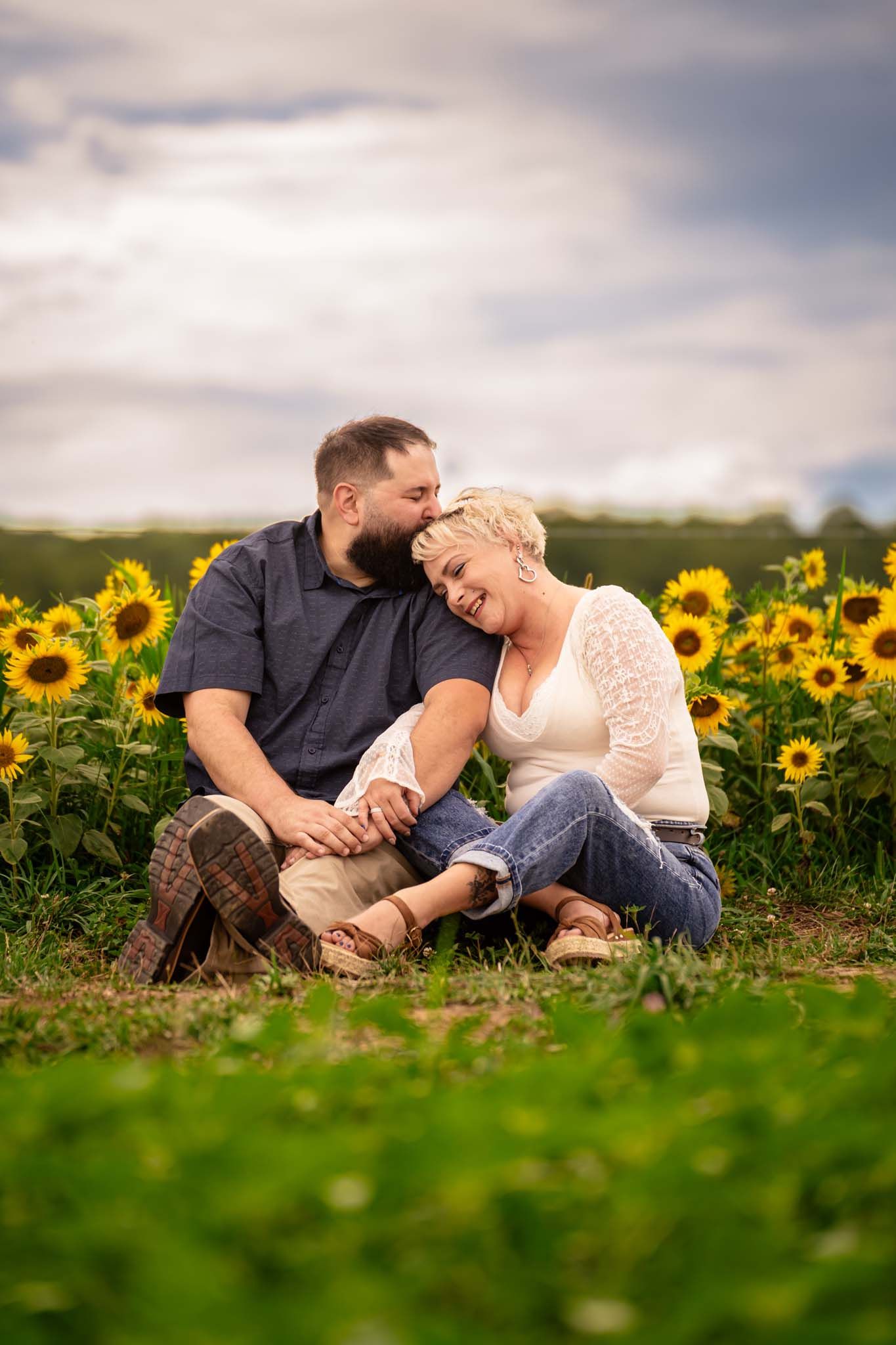 Couple sits romantically in sunflower field holding hands under dramatic cloudy sky.