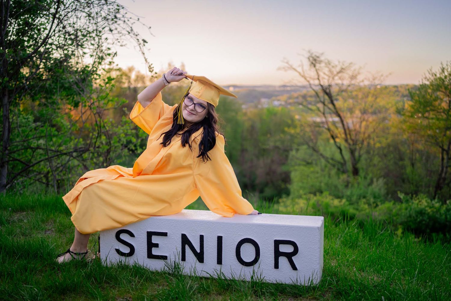 A senior girl in yellow cap and gown sits on a white SENIOR block outdoors at sunset.