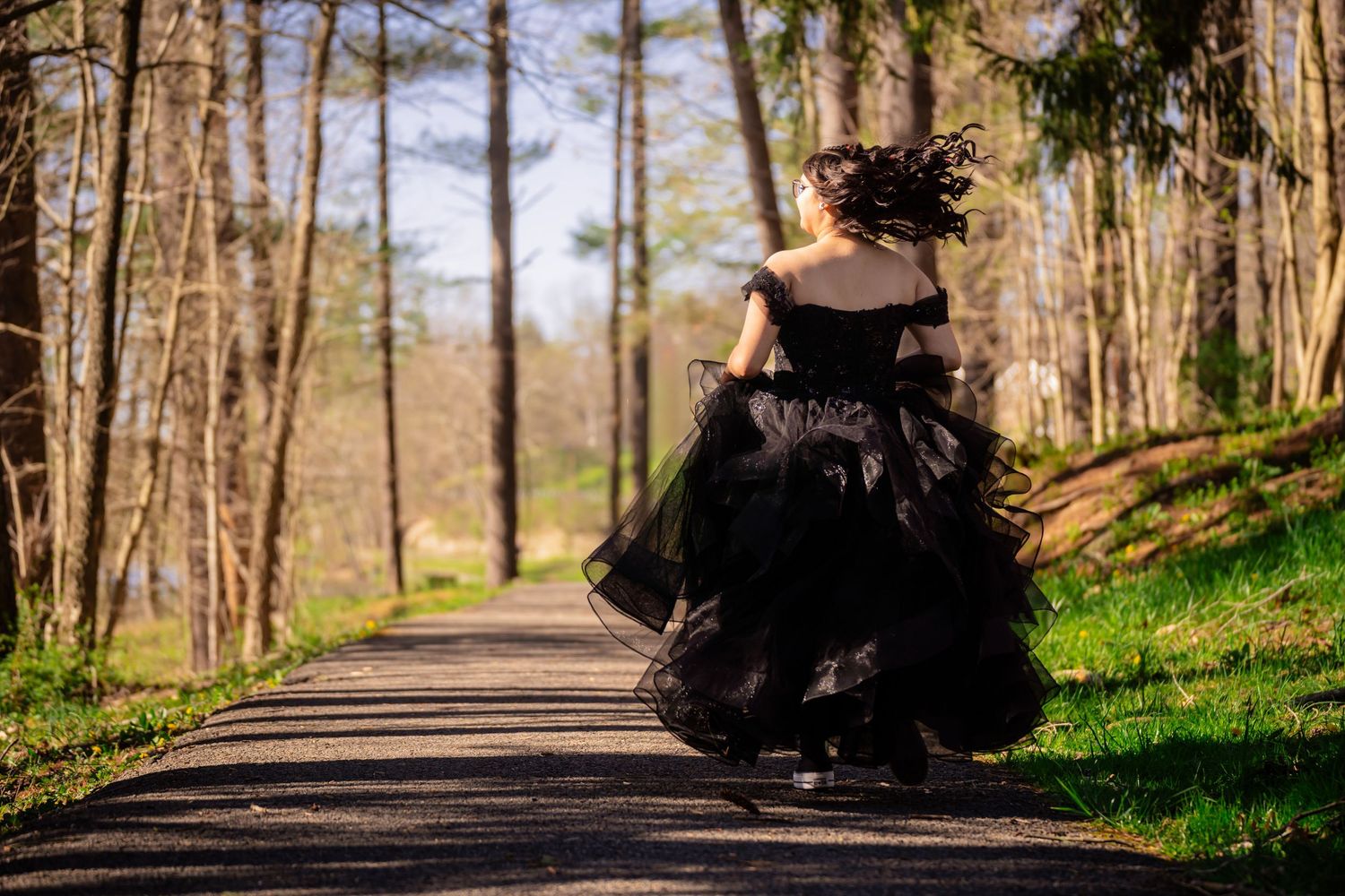 Woman in a flowing black ball gown walking away down a tree-lined path in sunlight.