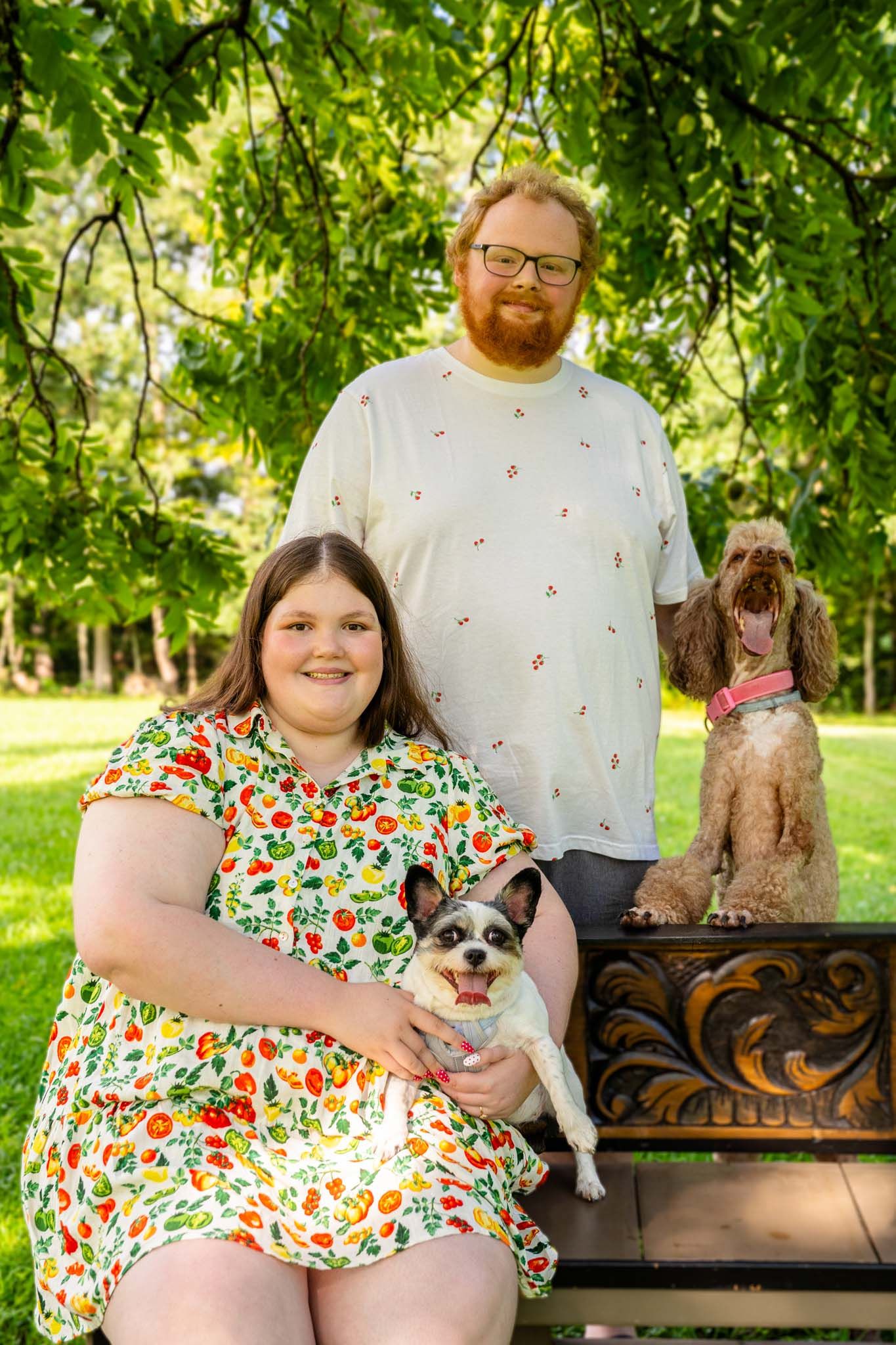 Woman in floral dress holds small dog on bench while bearded man and fluffy poodle pose in lush green park.
