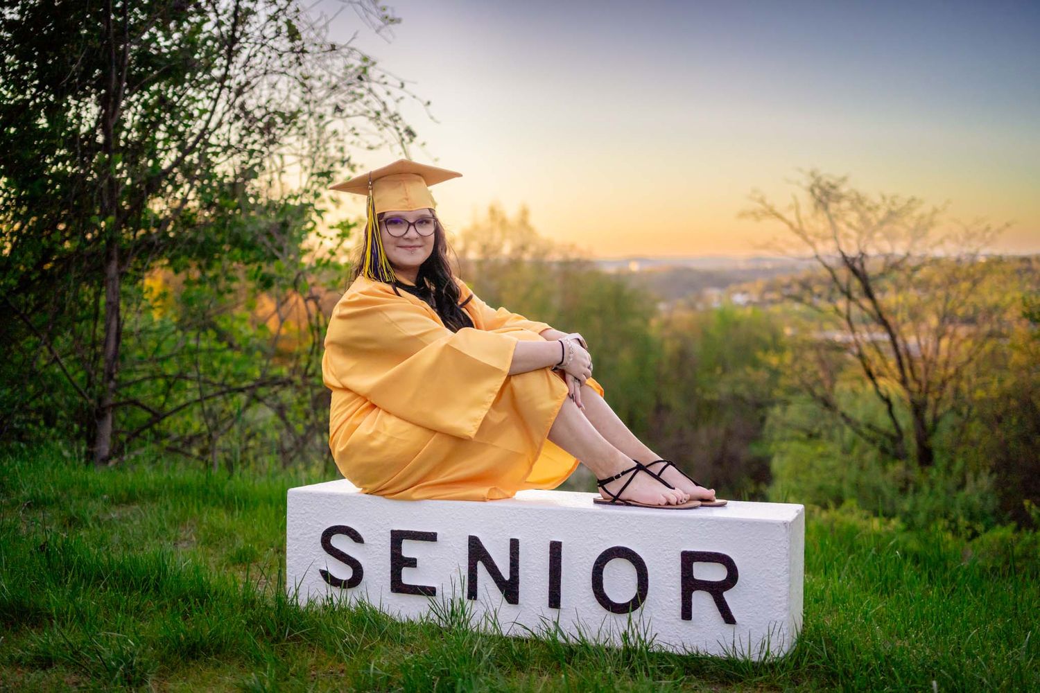 Female senior in gold cap and gown sitting on a white Senior block at golden hour outdoors.