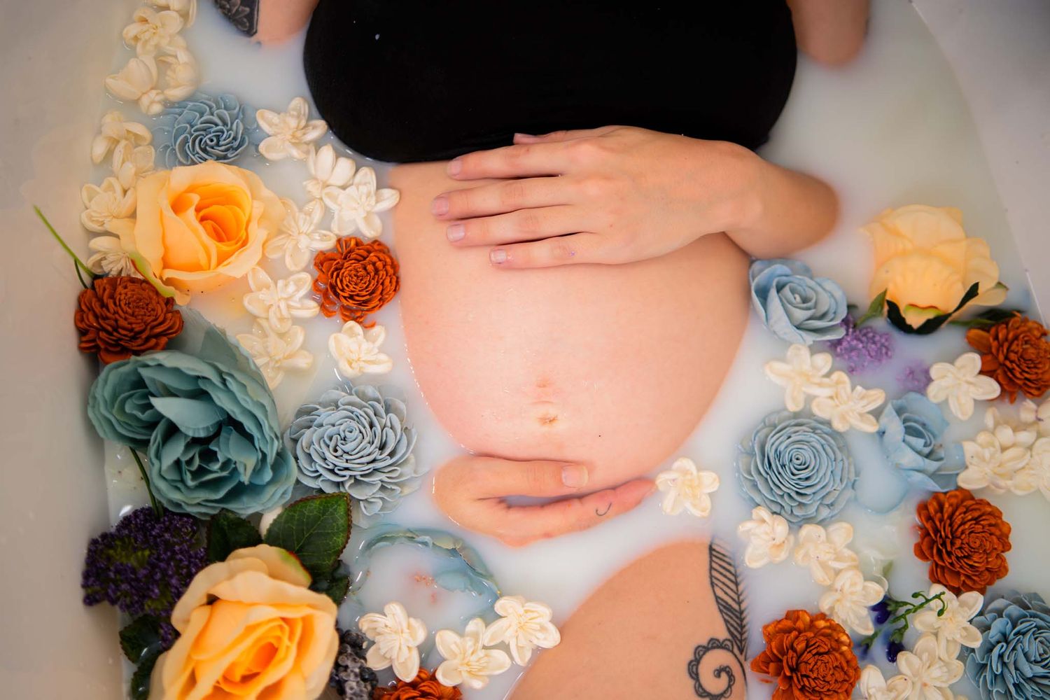 Close-up of pregnant belly cradled by hands in a milk bath with colorful flowers.