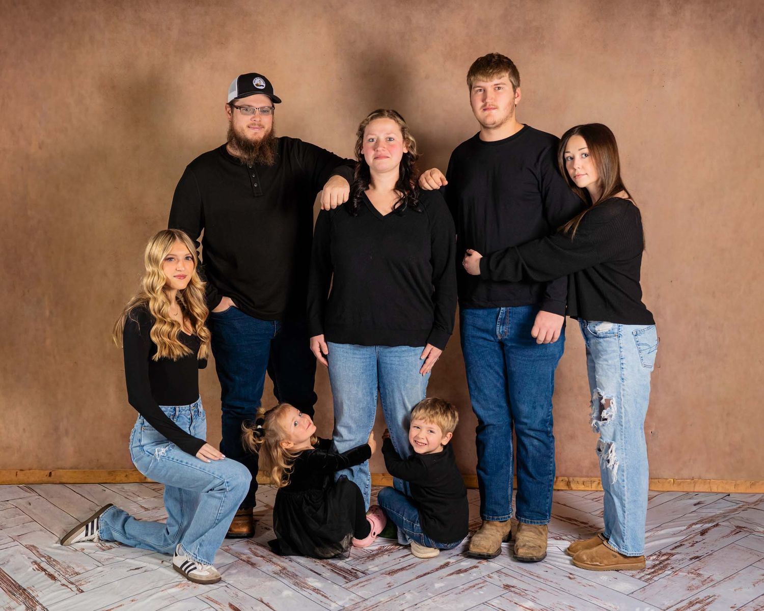 Family of seven posing for a professional portrait in matching black tops and jeans against a warm brown studio backdrop.