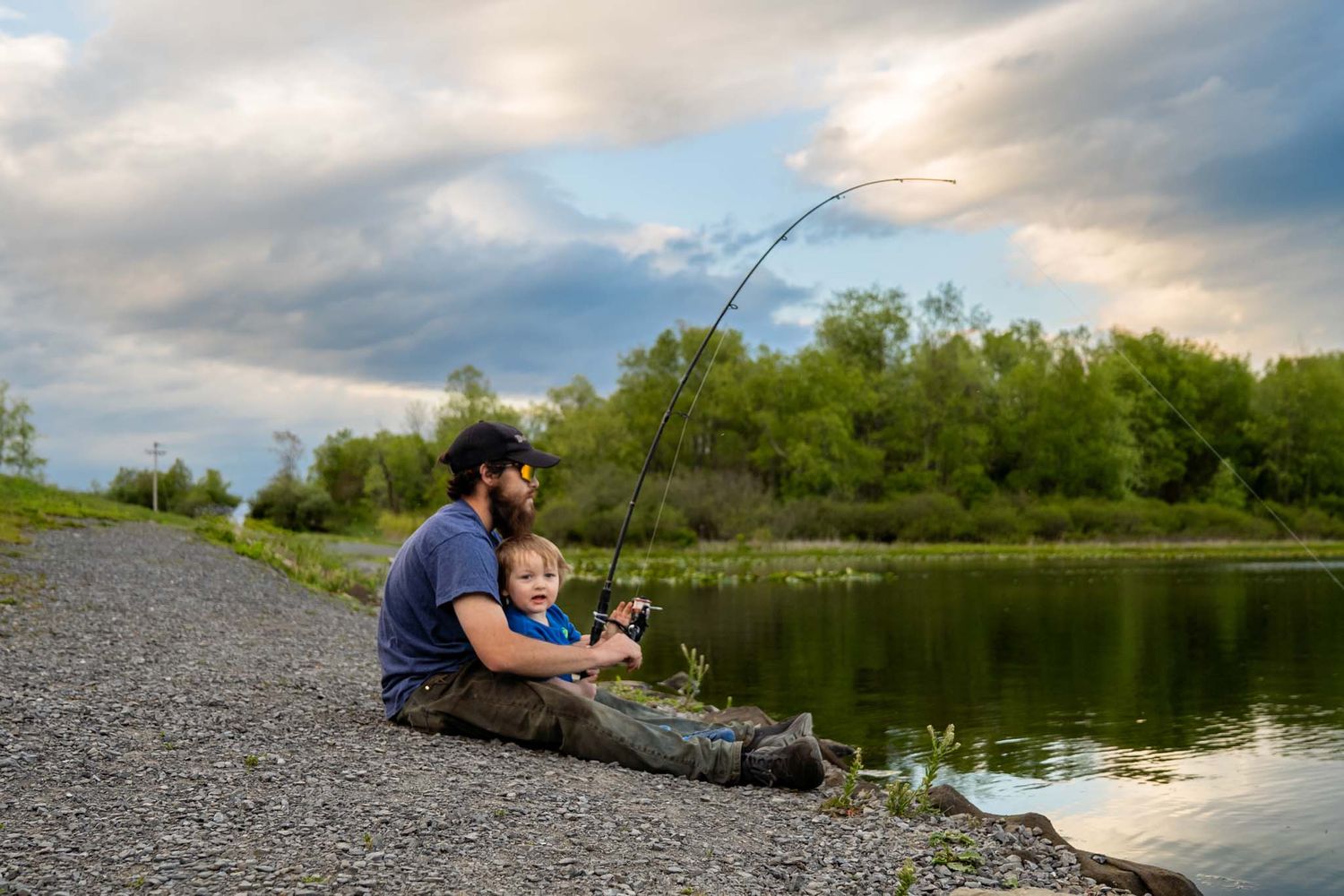 Father and young child sitting on rocky shore fishing together at a peaceful river with lush green trees and dramatic cloudy sky.
