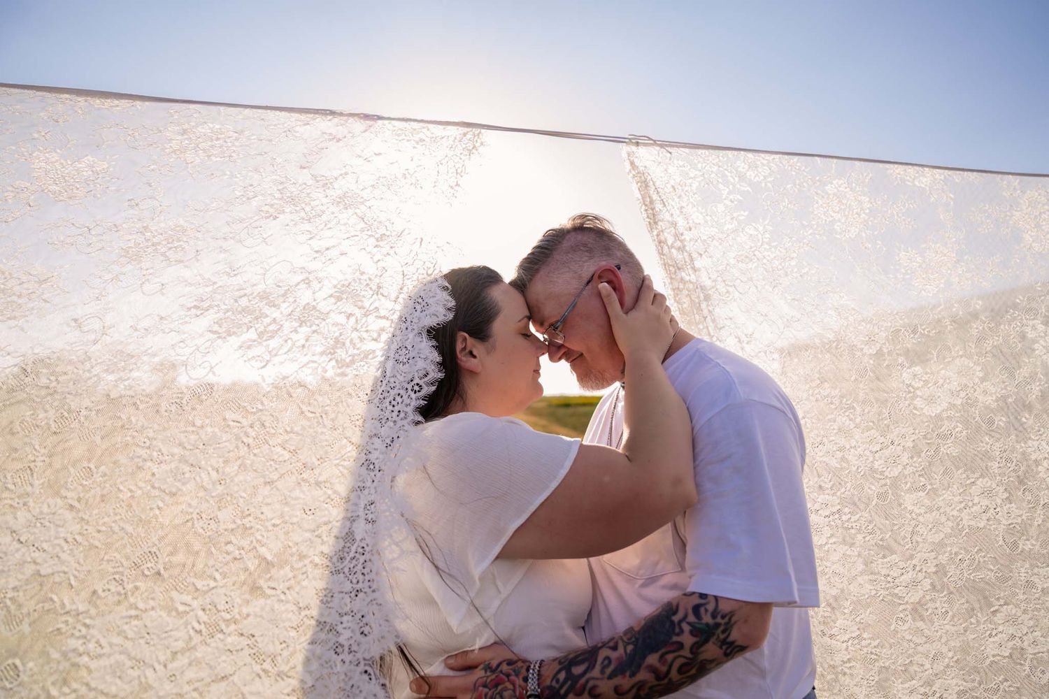 Romantic couple almost kissing under flowing white lace veil in bright sunlight outdoors.
