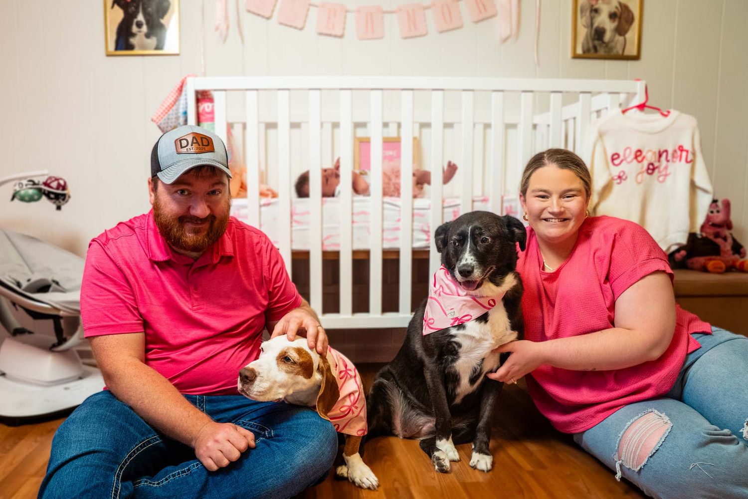 Happy new parents in pink shirts sitting on nursery floor with two family dogs beside white crib.