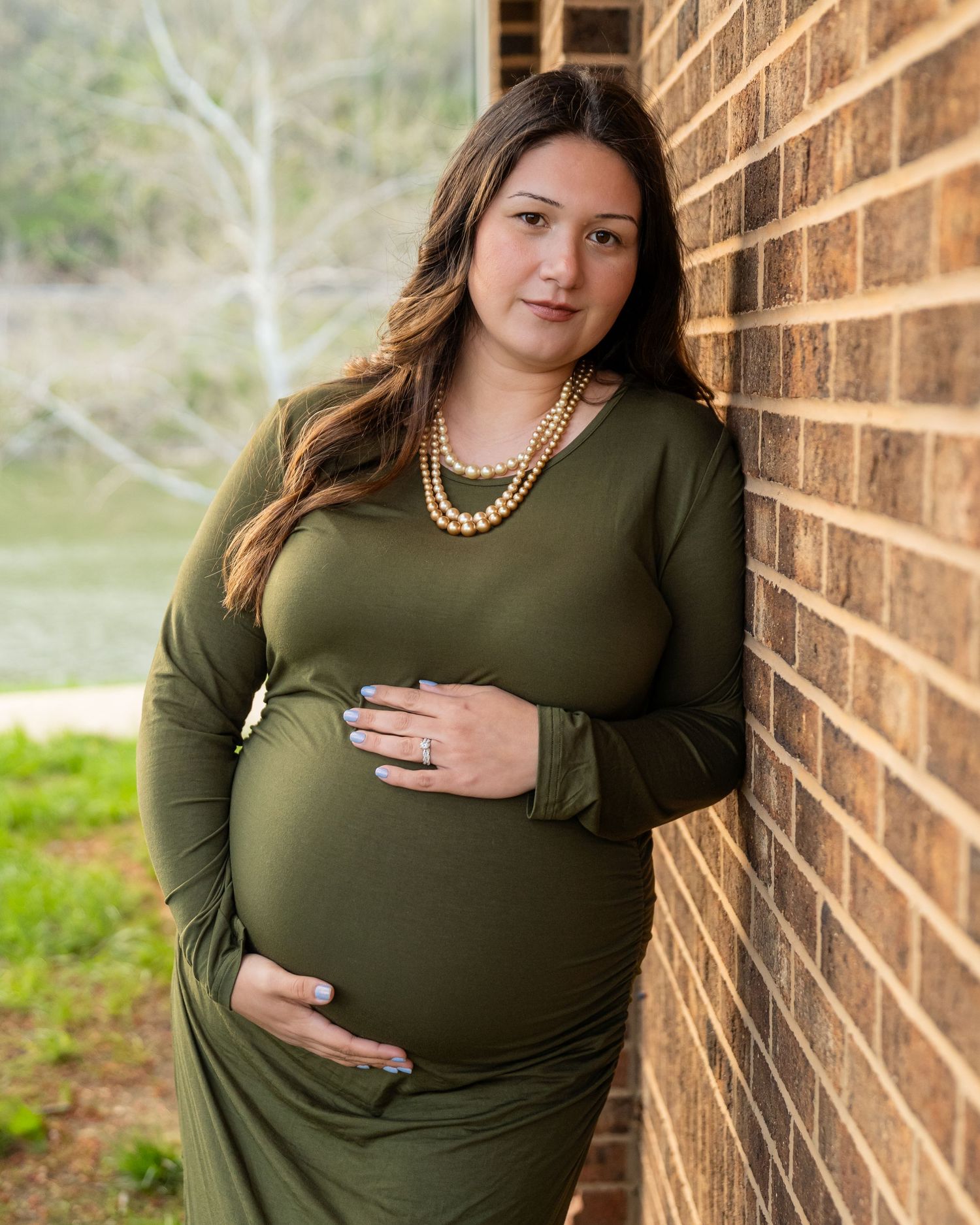 Pregnant woman in olive green dress cradles baby bump while leaning on brick wall.