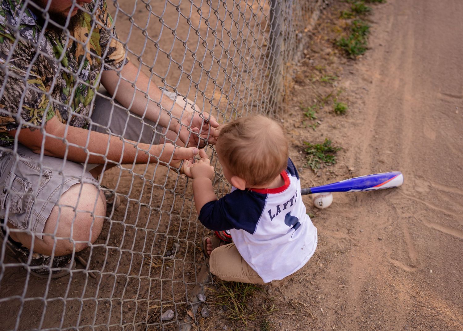 Toddler in baseball jersey sits by chain-link fence holding bat with ball nearby on dirt path.