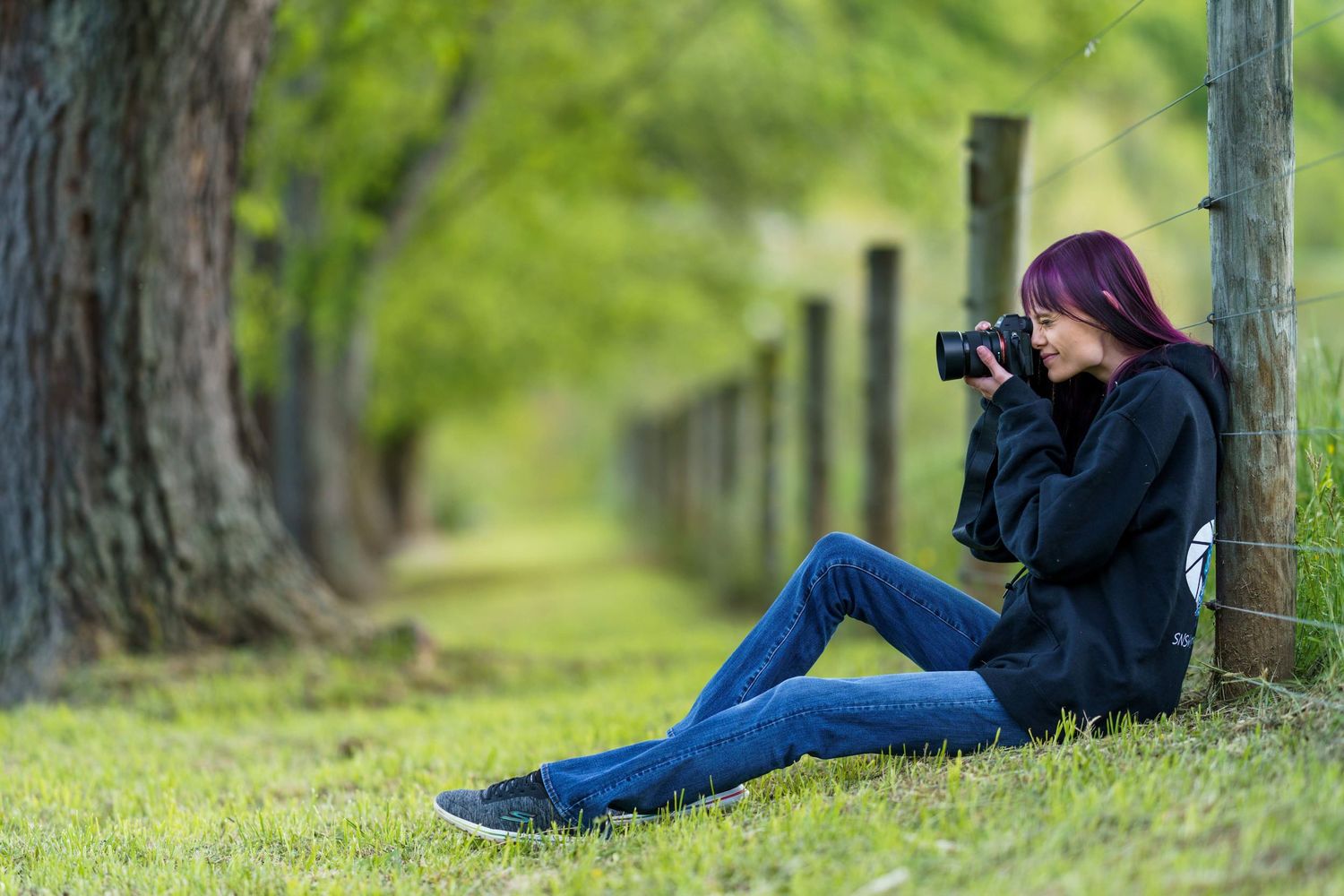 Woman with red hair sitting against fence post in tree-lined park, photographing nature with DSLR camera.