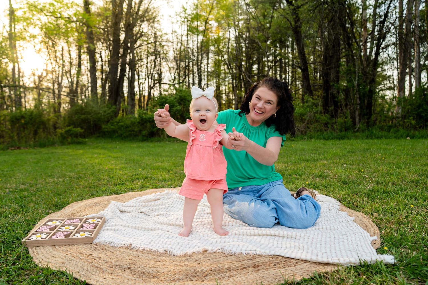 Mother helping baby girl take first steps on blanket in sunny green park during outdoor photo session.