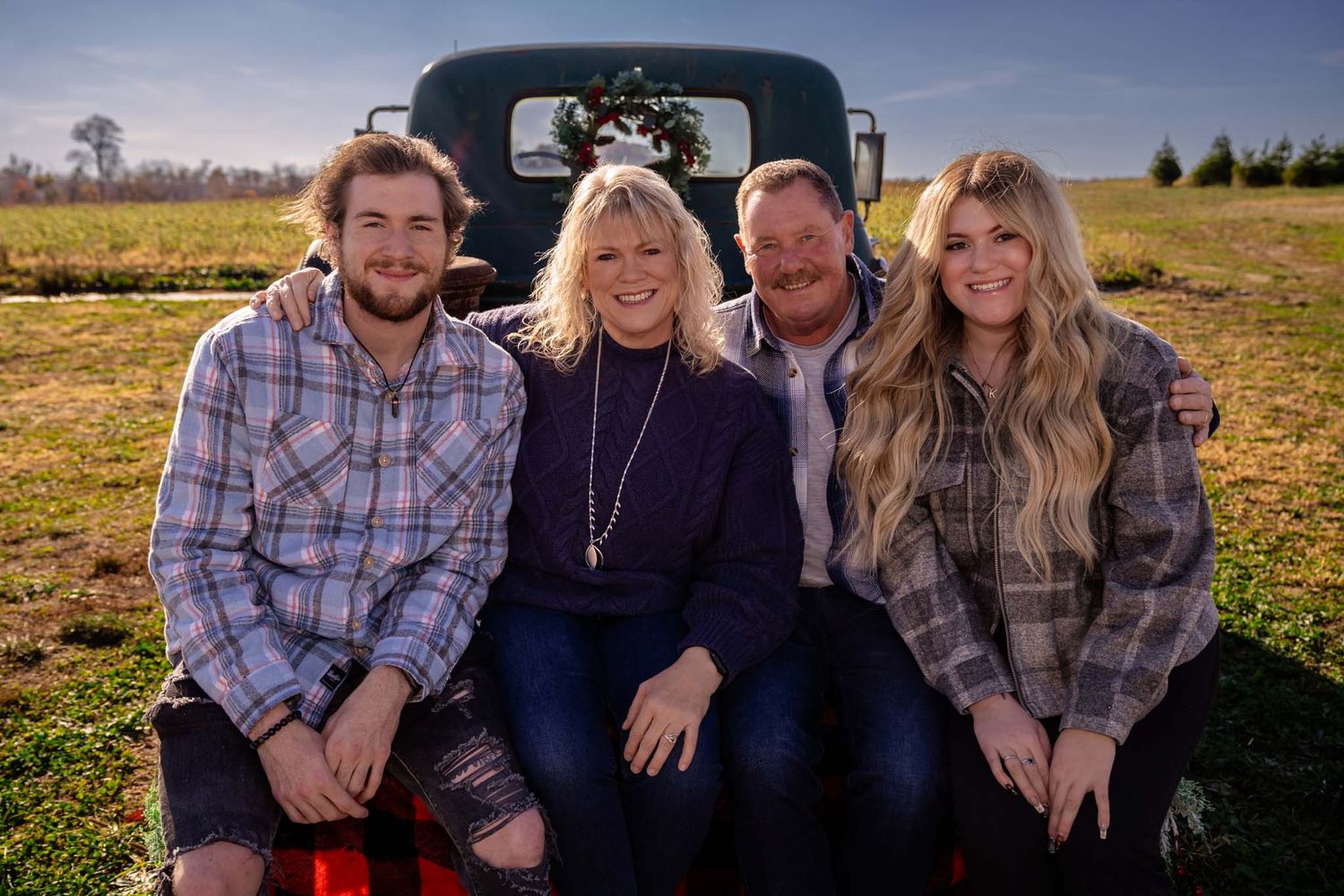 Family of four posing on the tailgate of a vintage truck decorated for Christmas on a farm.