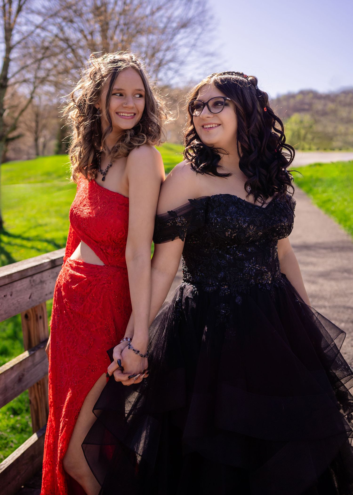 Two teenage girls in elegant prom dresses, one red and one black, holding hands on a bridge outdoors.