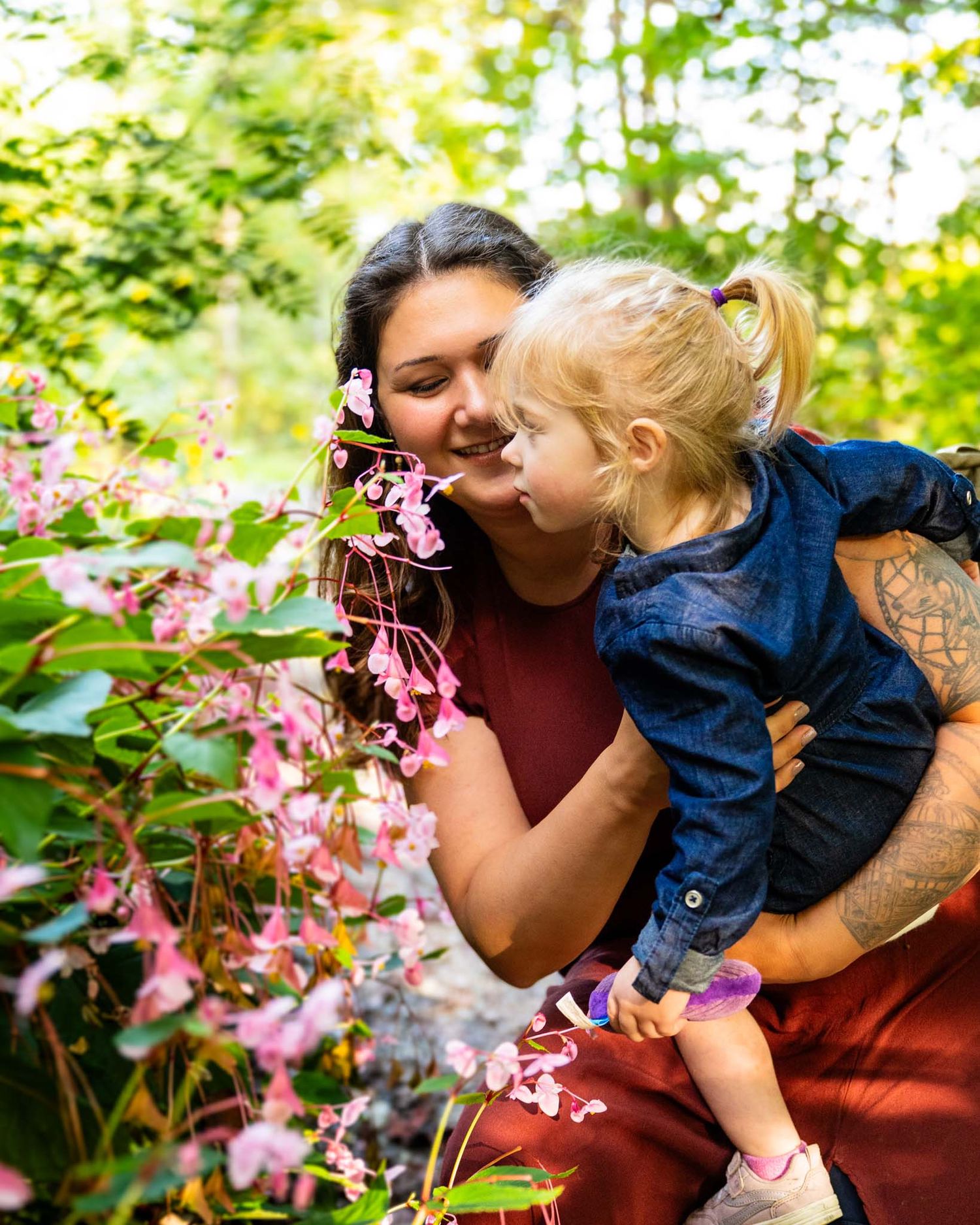 Mother holding toddler girl smelling pink flowers in sunlit garden setting outdoors.