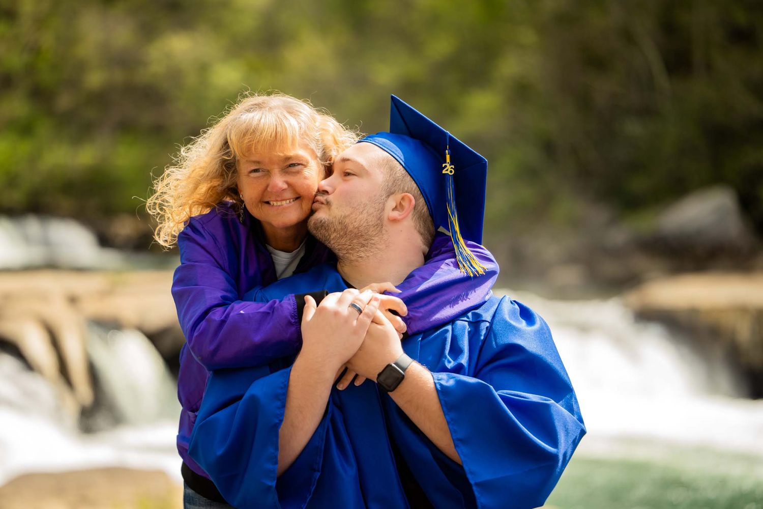 Graduate in blue cap and gown kissing a smiling woman on the cheek near a waterfall backdrop.
