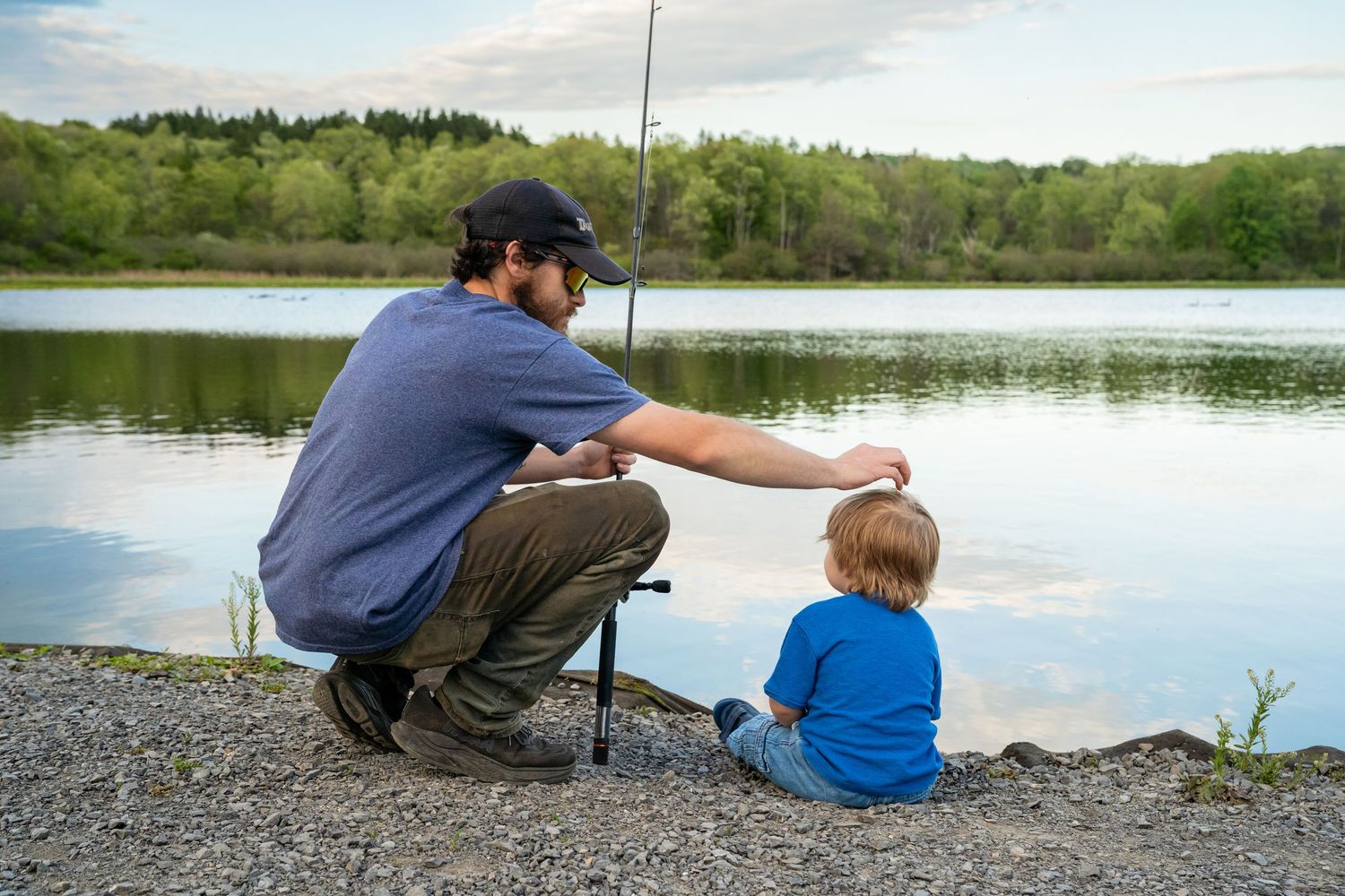 Father crouching beside young child fishing at a calm lakeside with green trees in background.