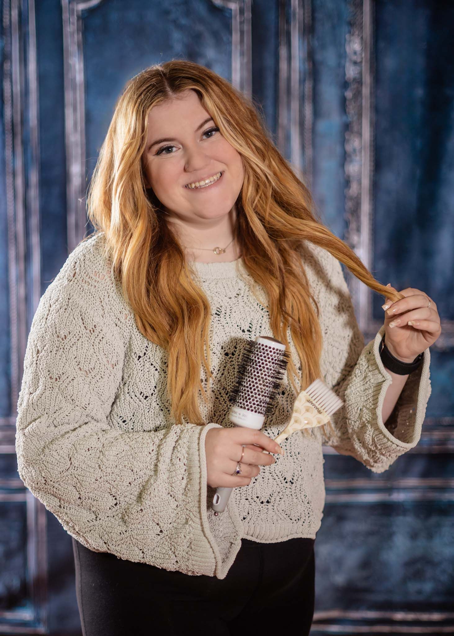 Smiling young woman with red hair holding round brush in cream knit sweater, blue backdrop.