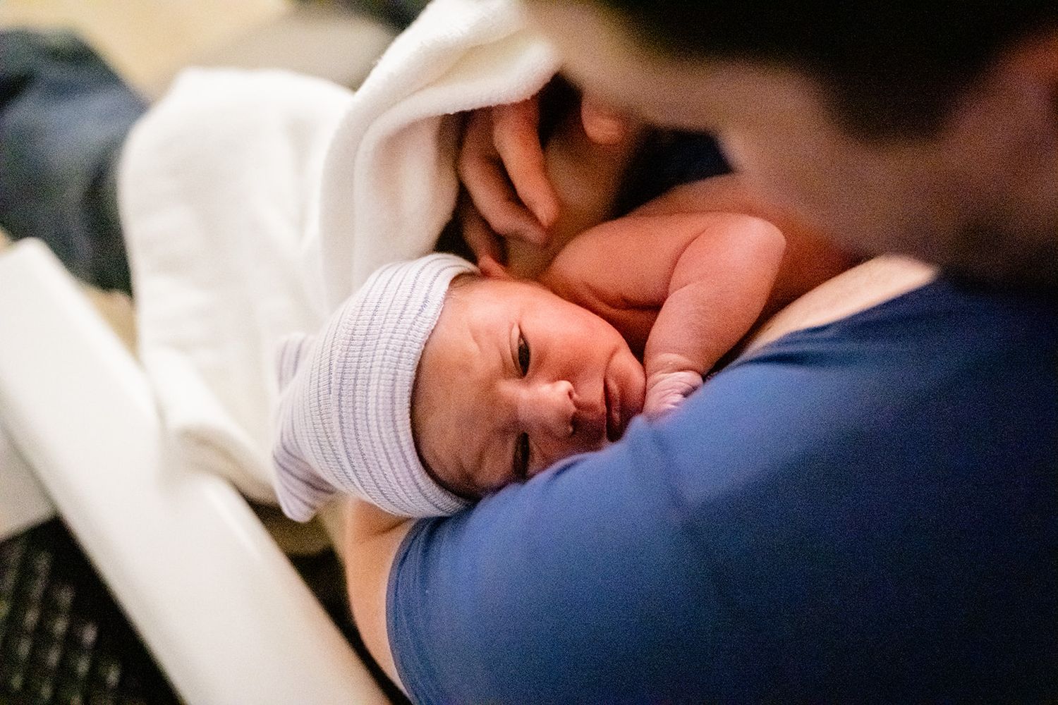 Father cradling a sleeping newborn baby wrapped in a white blanket wearing a striped hospital hat.