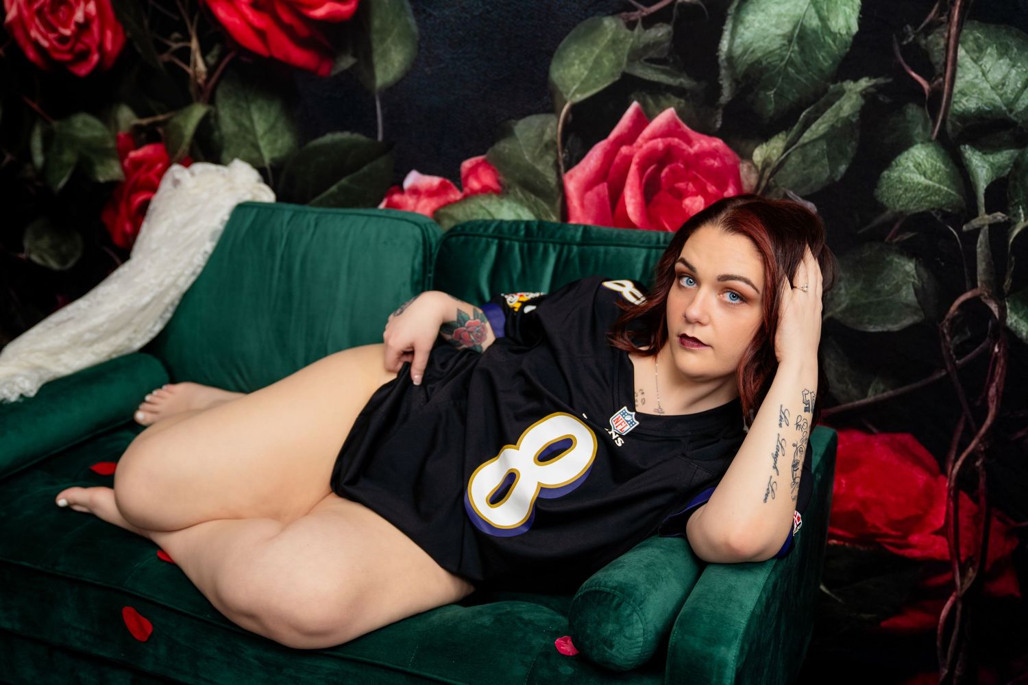 Woman in sports jersey reclining on green velvet sofa with floral rose wallpaper background.