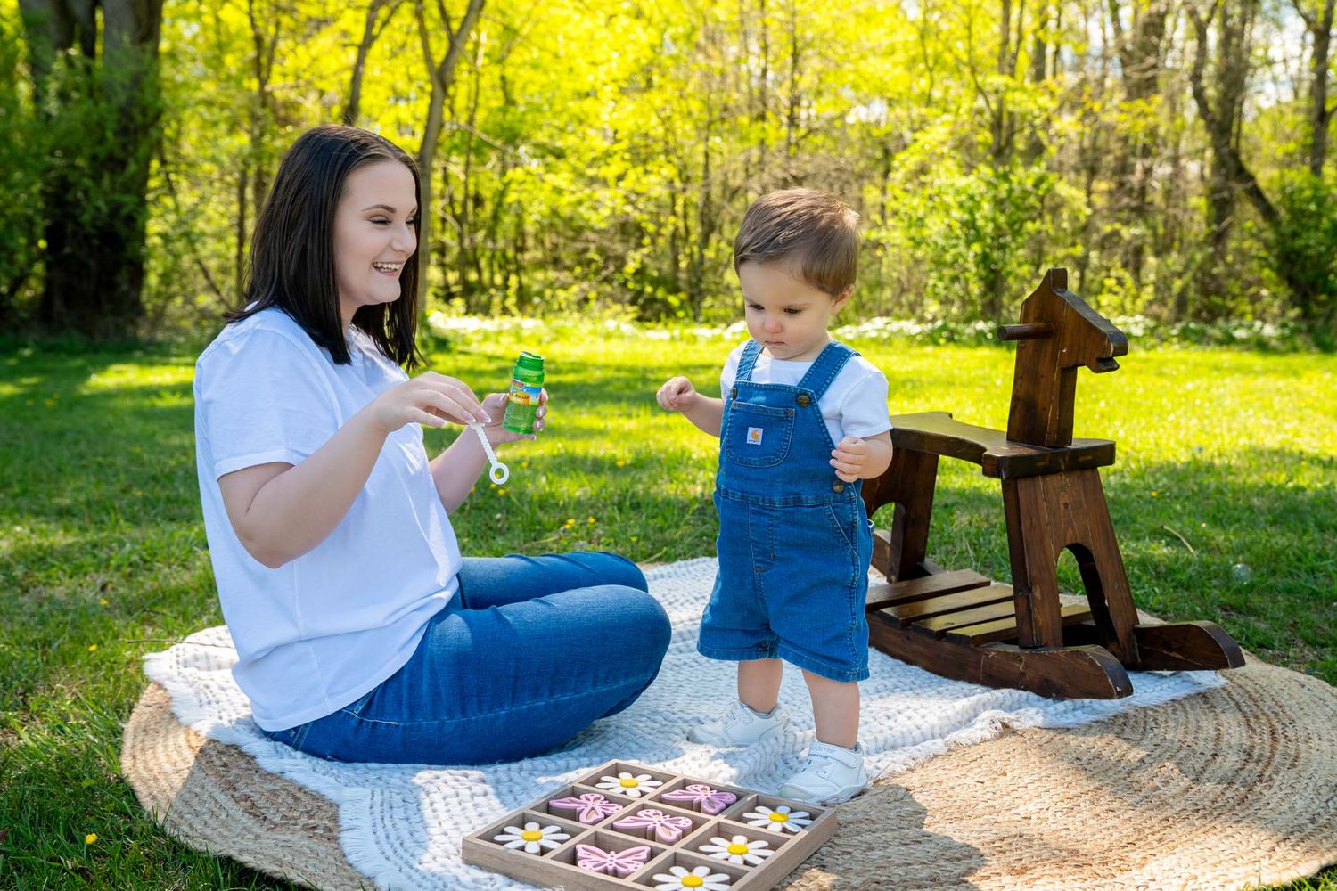 A mother and toddler play outdoors on a blanket in a sunny park with a wooden rocking horse nearby.